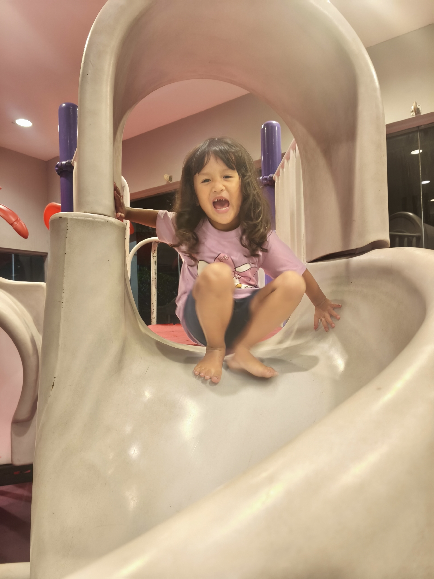 A young girl with long, dark, wavy hair is captured mid-slide on a plastic playground structure. She is wearing a light purple t-shirt with a cartoon character graphic and dark shorts. Her mouth is open in a joyful shout, and her eyes are wide with excitement. Her bare feet are positioned to control her descent, and her hands are gripping the sides of the slide.
The playground slide is made of a light-colored plastic, curving downwards. In the background, the ceiling is a pale pink, illuminated by soft lighting. A purple pole is visible to the left of the girl, and parts of a red structure can be seen peeking into the frame on the far left. The overall setting appears to be indoors, possibly in a play area or entertainment center, given the artificial lighting and the enclosed nature of the structure. The image conveys a sense of fun and energetic play, with the girl clearly enjoying her ride. The lighting suggests it is either daytime indoors or evening. There is no visible text in the image.