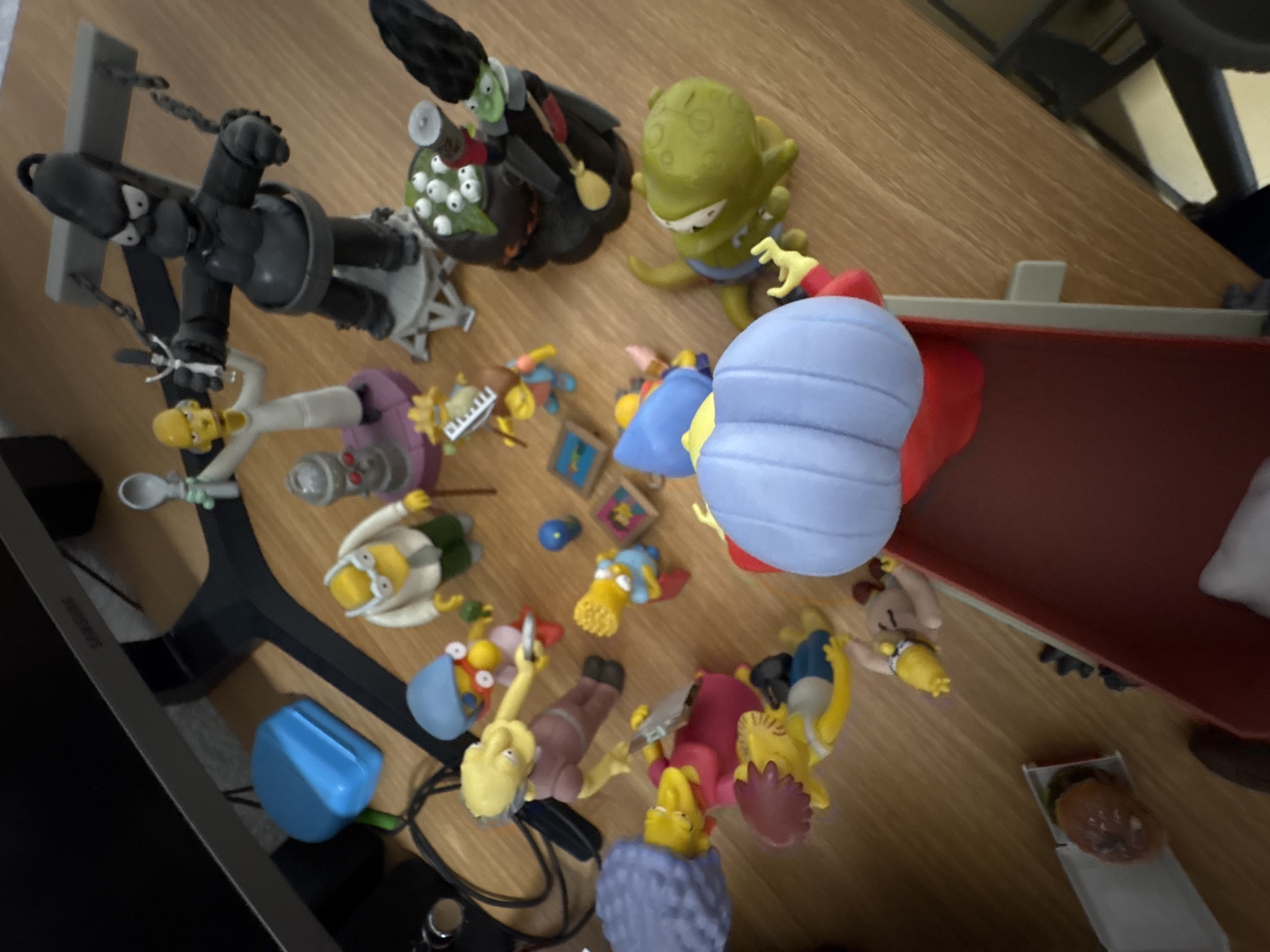 The image shows a collection of Simpson's figurines arranged on a wooden surface. The figurines include various characters like Homer, Bart, Lisa, Marge, and characters from the Treehouse of Horror episodes like the witch and Kang. A black Homer Simpson in a straitjacket is visible, as well as Maggie Simpson being pulled across the screen by a toy train carrying a hamburger. The figurines are arranged haphazardly, suggesting they may have been placed there for play or display.
The location in the city of Cuernavaca, Mexico is not discernible from the image. There is no identifiable landmarks in the image that would allow for such a determination.