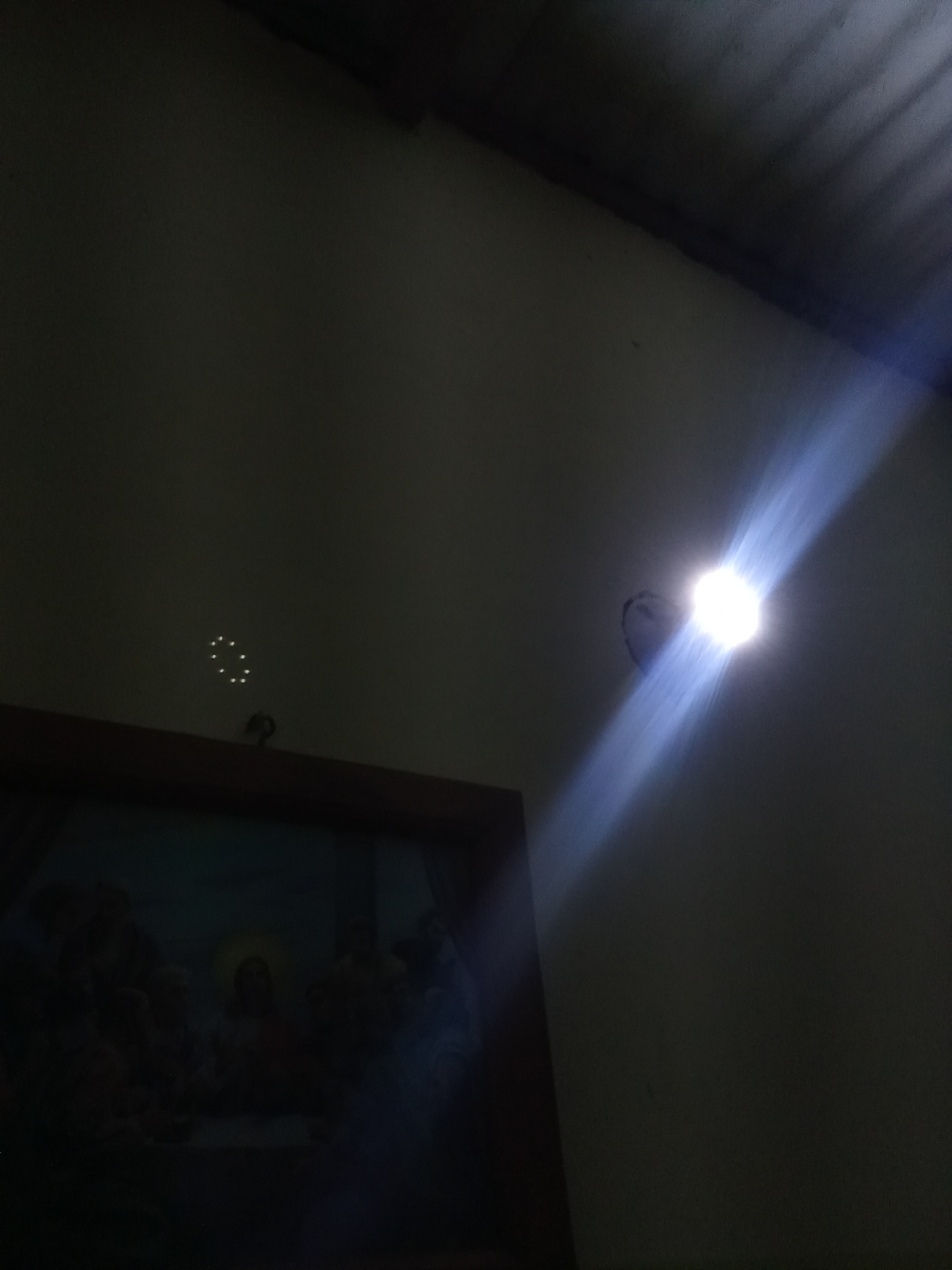 A dark, indoor scene shows a room with a framed picture on the lower left and a beam of light entering from a hole in the wall on the right. The light beam is bright and has a distinct ray-like appearance. Above the picture, there's a faint circular pattern of lighter spots on the wall. The upper portion of the image suggests a ceiling. The location in Juan Griego, Venezuela, is not directly discernible from the visual elements.