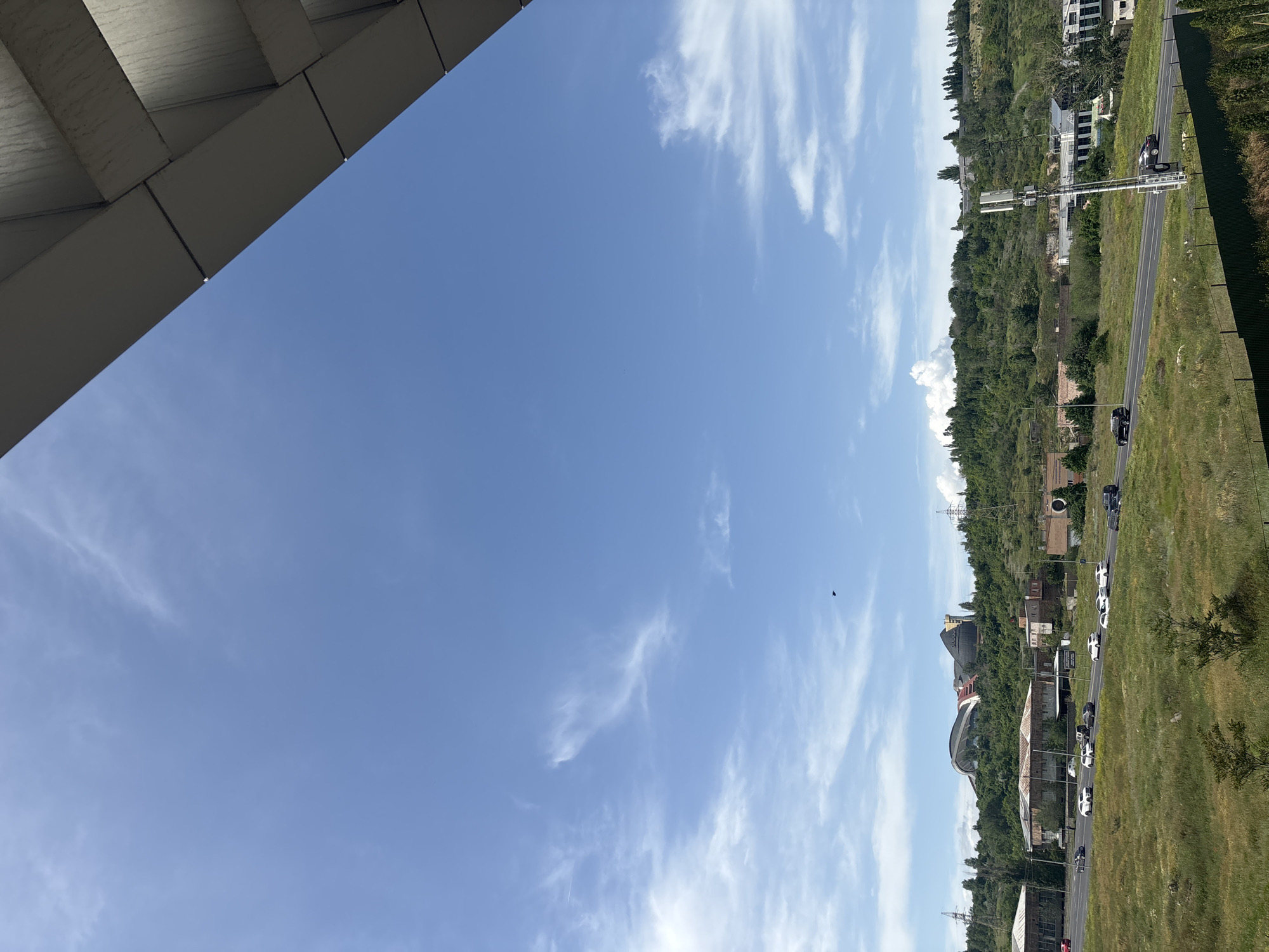 The image captures a scene in Yerevan, Armenia, featuring a clear blue sky with scattered cirrus clouds. The lower right of the frame shows a landscape including a road with vehicle traffic, green fields, trees, and buildings. A large architectural structure is visible. The upper left corner contains a section of a gray concrete or metal structure, possibly part of a building or bridge. The image is rotated 90 degrees counterclockwise.
