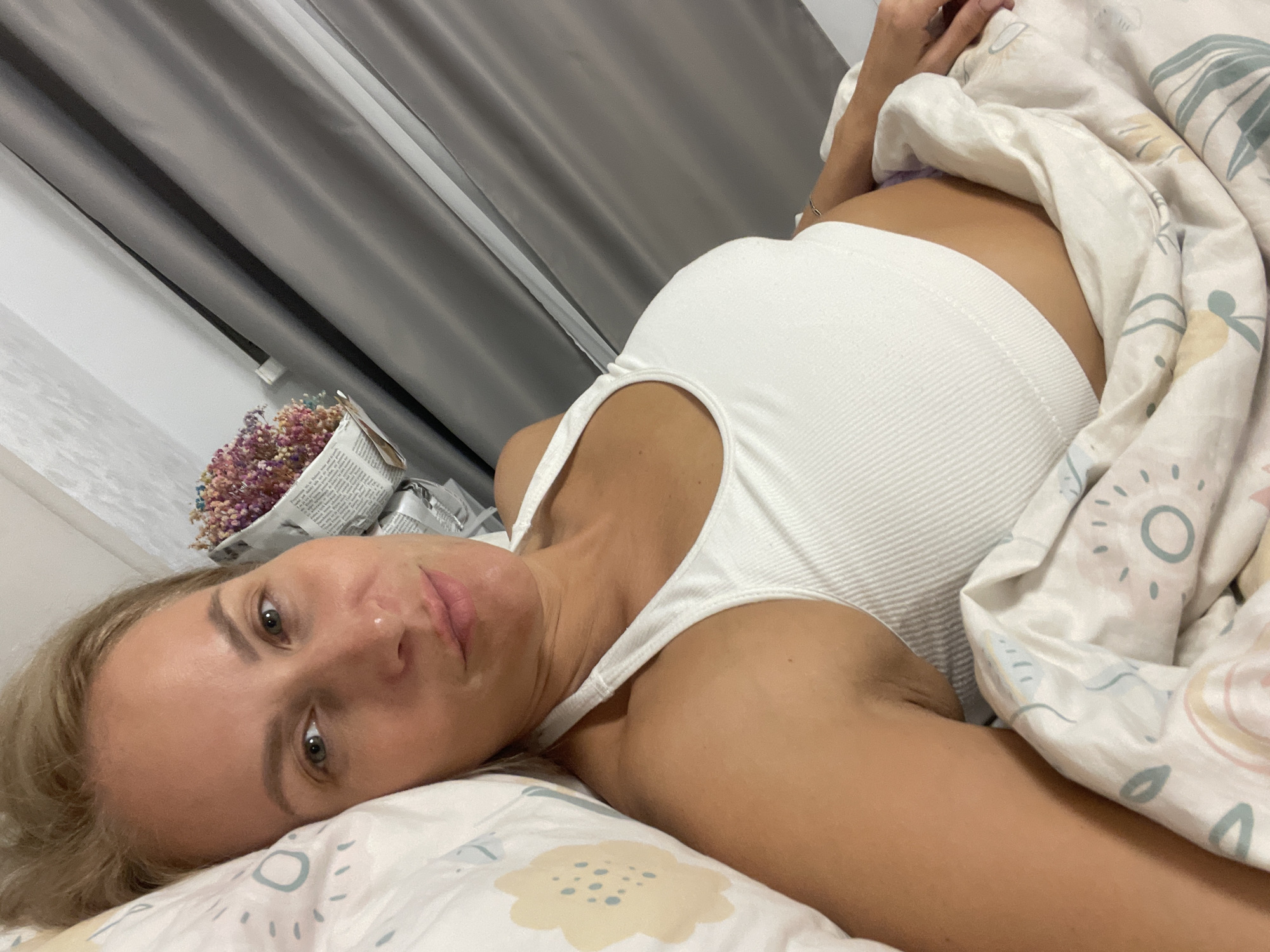 A woman with blonde hair lies on a bed covered with a patterned white blanket. She is wearing a white top. Her head rests on a pillow with a yellow flower pattern. Beside her, on a bedside table, is a bouquet of flowers wrapped in newspaper. Gray curtains are in the background. The location of Baranovka, Russia, is not discernible from the provided image.
