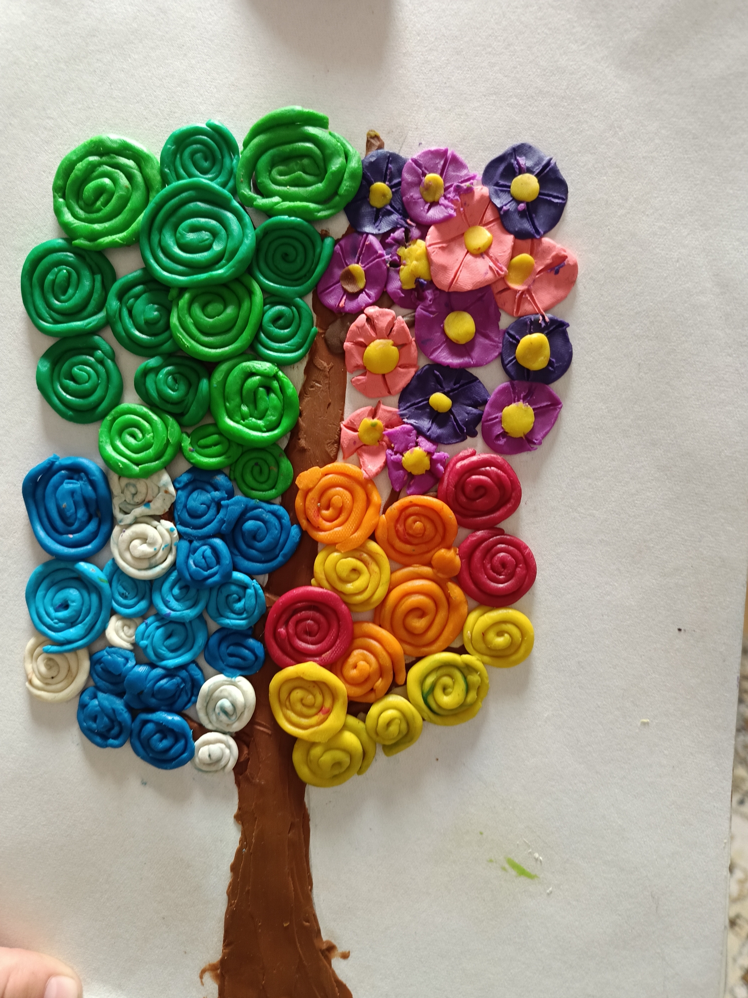 A child's artwork depicts a four-season tree created with modeling clay on white paper. The tree trunk is brown. The four quadrants of the tree's canopy represent the seasons: spring (blue and white), summer (green), autumn (orange and red), and winter (flowers). The location in Chaikovsky, Russia is unknown.