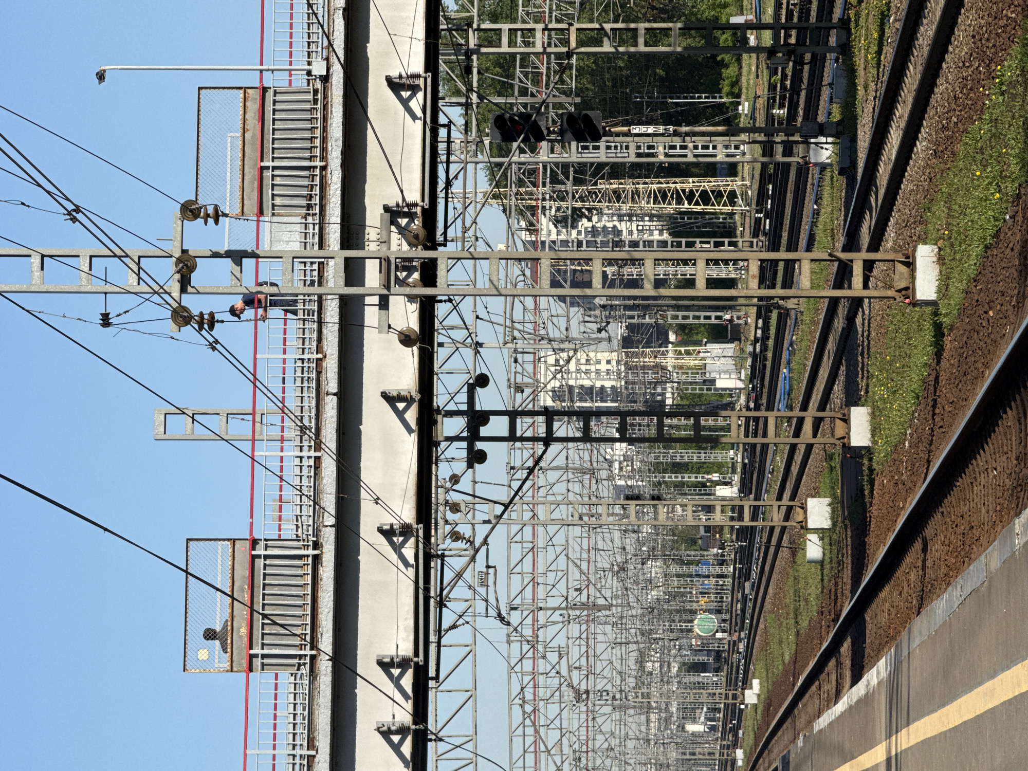 The image depicts a railway scene in Moscow, Russia. The view is taken from a station platform, marked by a yellow line, looking along several sets of railway tracks. Above the tracks, there is an extensive network of overhead power lines and metal support structures. In the distance, building and trees are visible. On the left side of the image, there is a building with staircases and platforms enclosed by wire mesh. A person in a blue shirt and pants appears to be working on or inspecting equipment on one of these platforms.