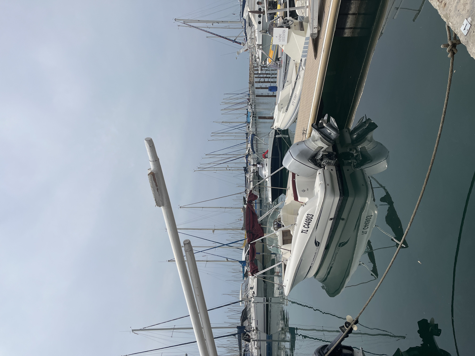 This image shows a marina in Palavas-les-Flots, France. In the foreground, a white speedboat with the registration TL C44903 is docked. Its reflection is visible in the dark water. Behind it, other boats, including sailboats and a small rigid inflatable boat with the registration STE 63881, are moored. In the background, a long row of sailboats with tall masts stretches into the distance. The sky is overcast and the water is calm. A white pipe, possibly part of a dock structure, is angled across the upper portion of the image.
