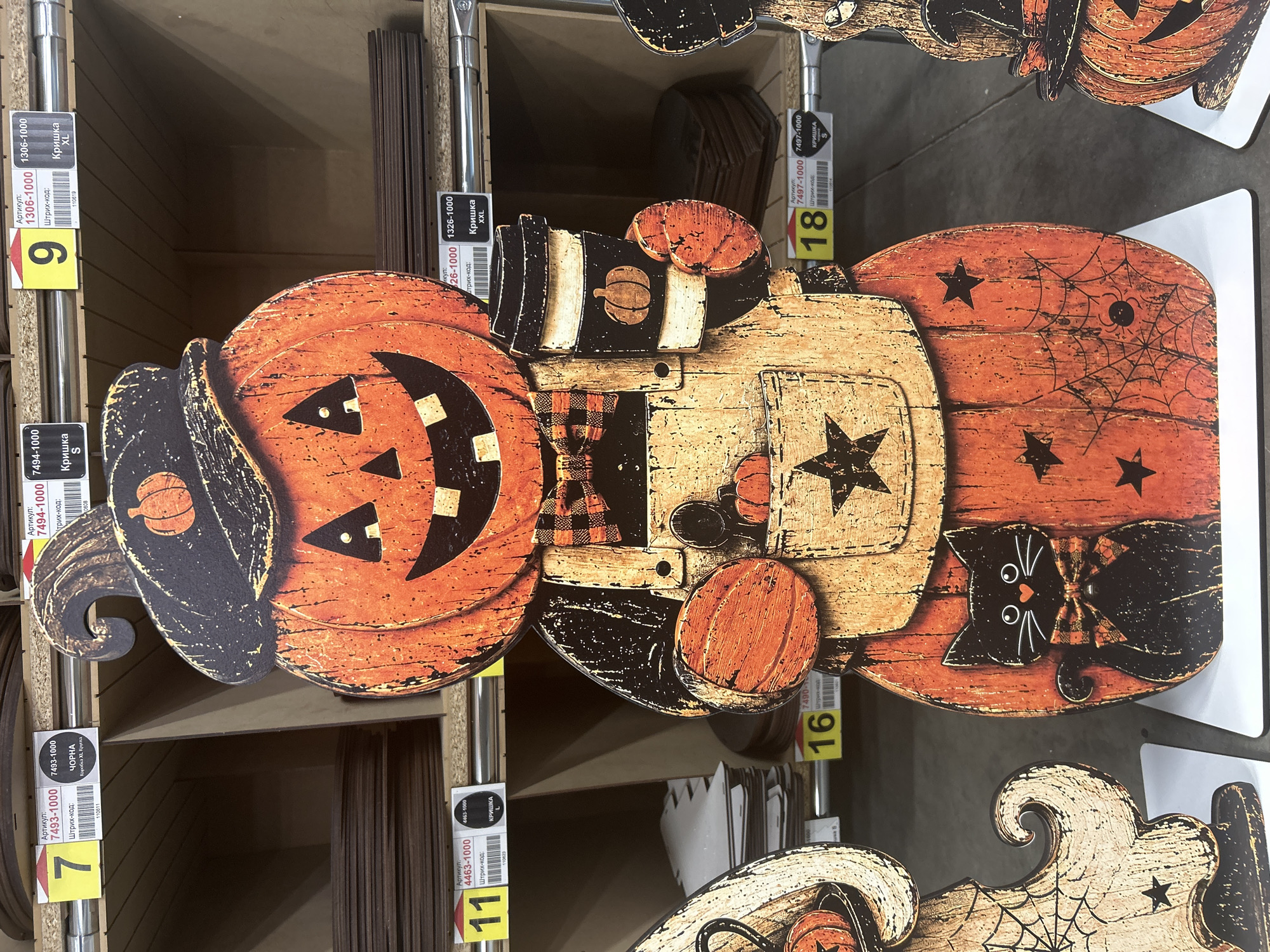 The image shows a festive Halloween-themed cardboard cutout of a smiling jack-o'-lantern character wearing a hat and an apron. The character is holding a coffee cup in one hand and a small pumpkin in the other. A black cat with a bow tie is depicted on the lower body of the cutout. The cutout is positioned in what appears to be a retail environment, with shelves of products visible in the background. Yellow price tags with Cyrillic text are attached to the shelves. The scene is brightly lit, highlighting the colors and details of the Halloween decoration. The location is identified as Ternopil, Ukraine, suggesting this is a retail display within that city.