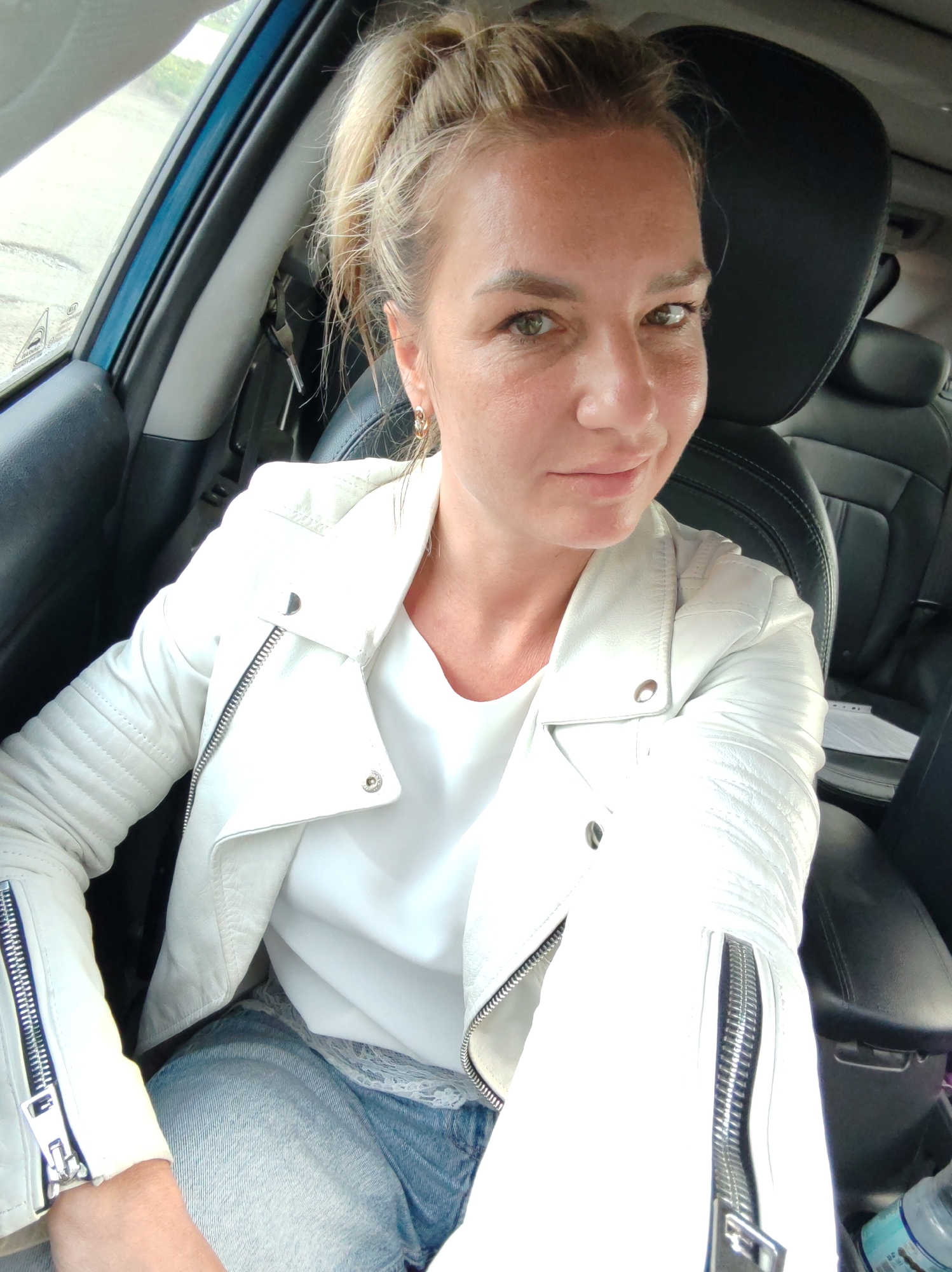A woman with blonde hair pulled into a ponytail, wearing a white leather jacket over a white top and blue jeans, is sitting in the driver's seat of a car. She is looking at the camera and smiling slightly. The car's interior, including the black leather seats and a portion of the dashboard, is visible. The scene is likely indoors or in a shaded area, as indicated by the lighting. There are no specific landmarks or environmental cues that would place the image in Petropavlovsk-Kamchatsky, Russia, or any other city.