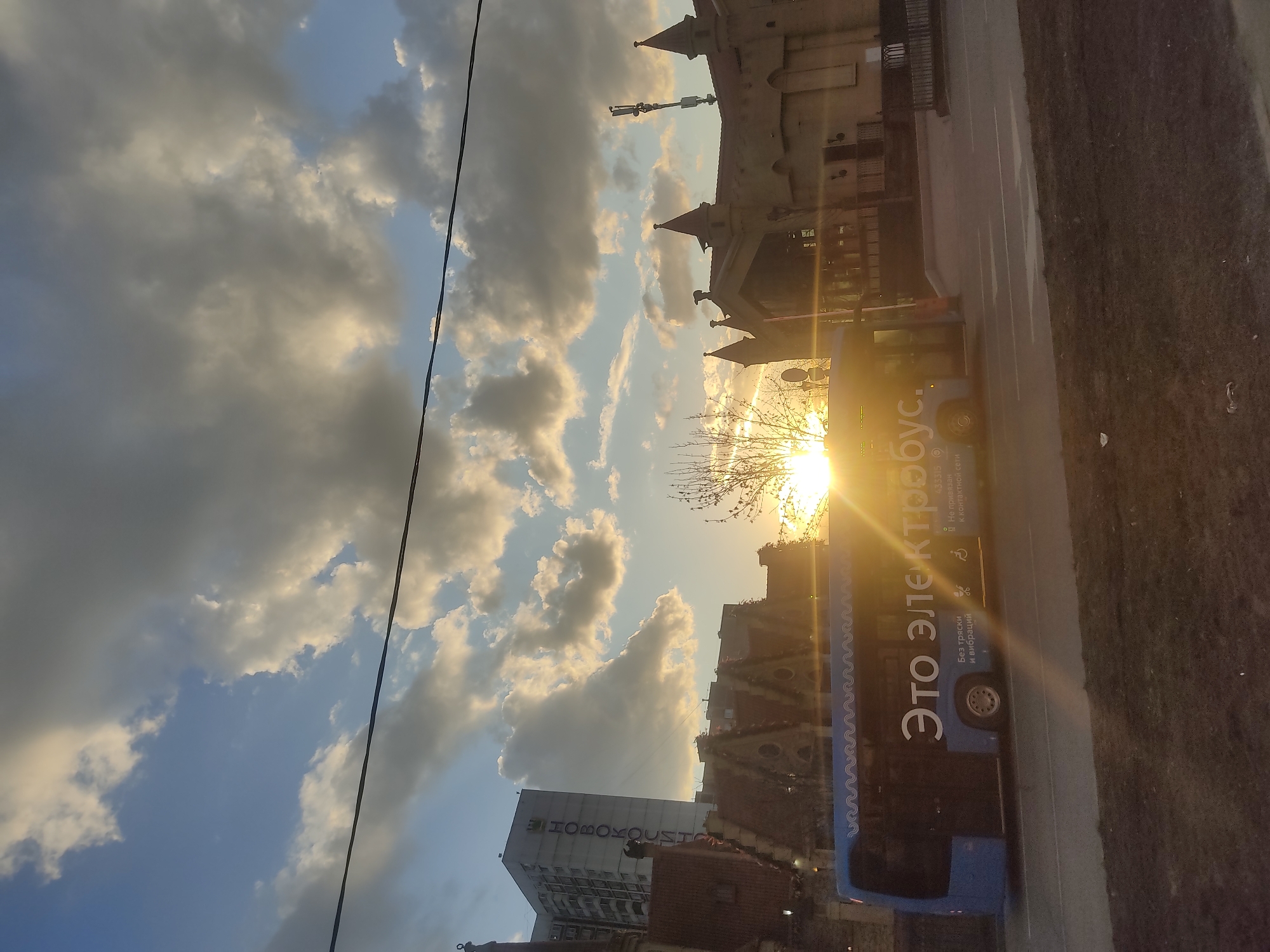 The image shows a bus in Moscow, Russia, with a building in the background. The bus has the words "Это электробус" written on it, which translates to "This is an electric bus". The sky is visible with clouds and the sun shining through. The image appears to be taken at sunset or sunrise.