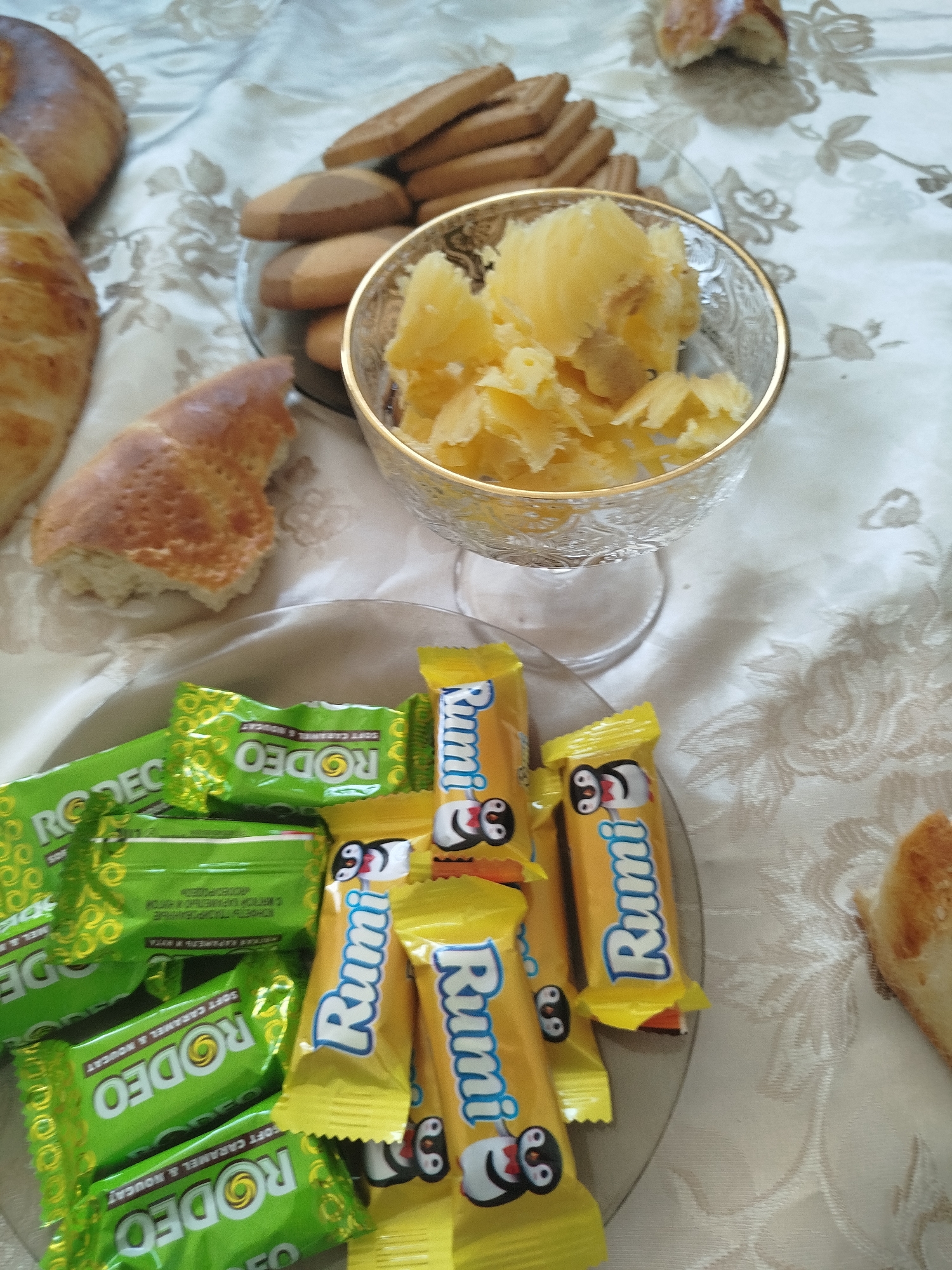 The image depicts a spread of food items on a tablecloth with a floral pattern. In the foreground, there are several wrapped candies, some in green packaging labeled "RODEO" and others in yellow packaging labeled "Rumi" with cartoon penguin characters. A plate with more of these candies is visible behind the initial layer. To the left, there are pieces of bread, one appearing to be a larger loaf and another a smaller, torn piece with a golden-brown crust. In the background, a glass bowl contains a yellow, shredded substance, possibly fruit or cheese. There are also several flat, rectangular cookies stacked on a plate. The setting appears to be a domestic environment, potentially a table laid out for a meal or snack. No people are visible in the image. The location is identified as Жаңы-Айыл, Kyrgyzstan.