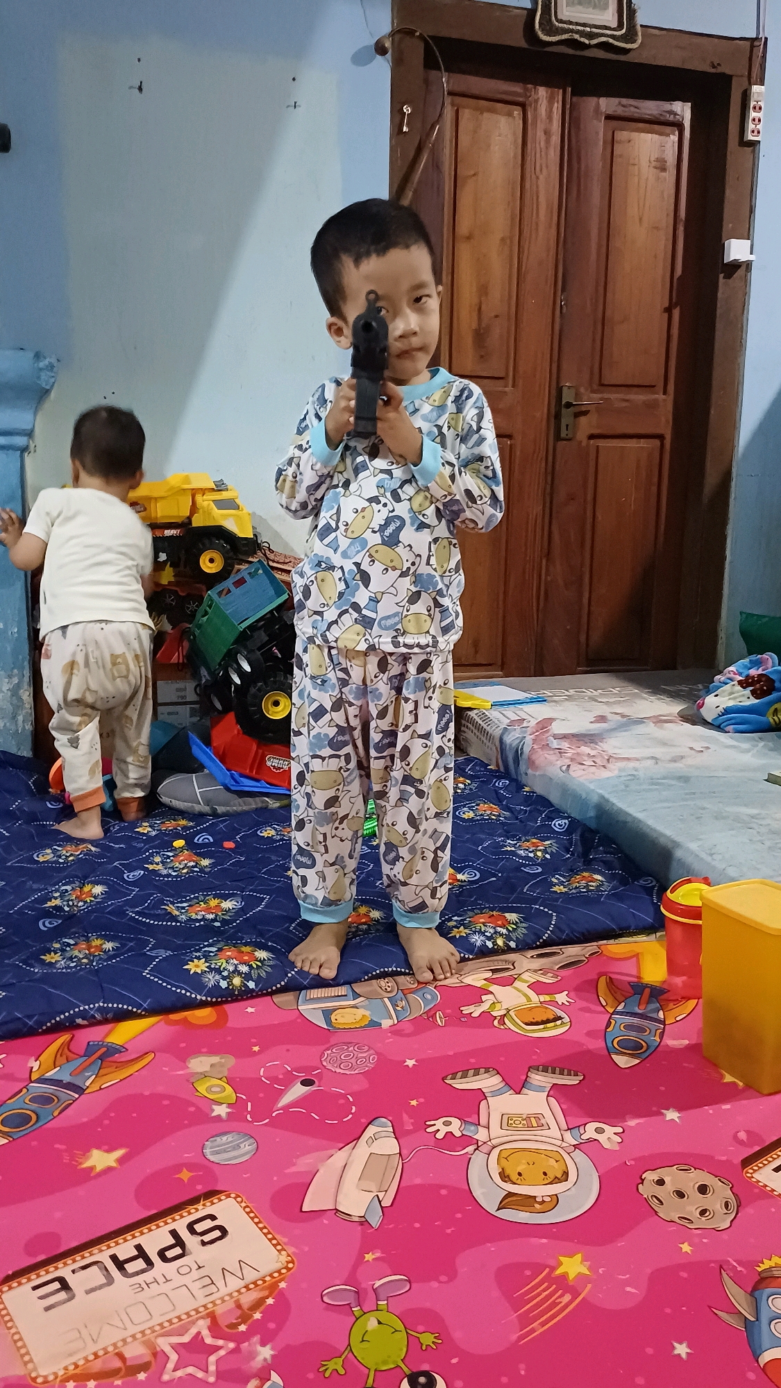 A young boy, wearing pajamas with a cow pattern, stands in the center of a room, holding a toy gun. His gaze is directed towards the camera. Beside him, another young child, facing away from the camera, is playing with a stack of large toy trucks. The scene appears to be a residential interior, possibly a playroom, with a wooden door and blue walls. The floor is covered with two large mats, one with a blue starry pattern and the other a vibrant pink with space-themed illustrations. In the background, a mattress is visible, possibly used for playing or sleeping. The overall environment suggests a domestic setting in Wonokerso, Indonesia, where children are engaged in play.