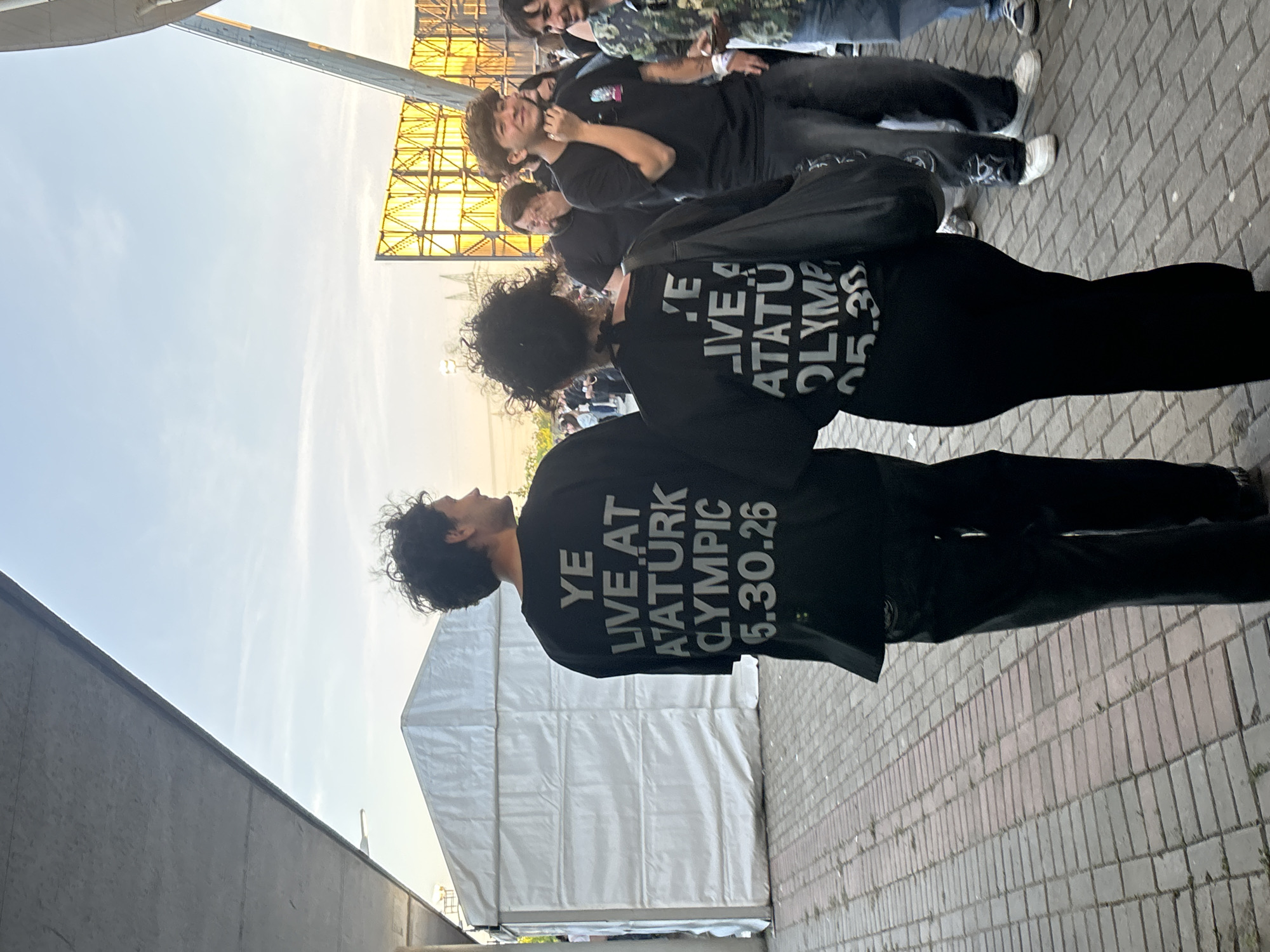 A group of people are standing on a brick sidewalk in Başakşehir, Turkey. Two people in the foreground wear black long-sleeved shirts with the text "YE LIVE AT ATATURK OLYMPIC 5.30.26" printed in white on the back. A white tent structure is behind them. Behind these two are other individuals; a tall billboard structure appears in the background.