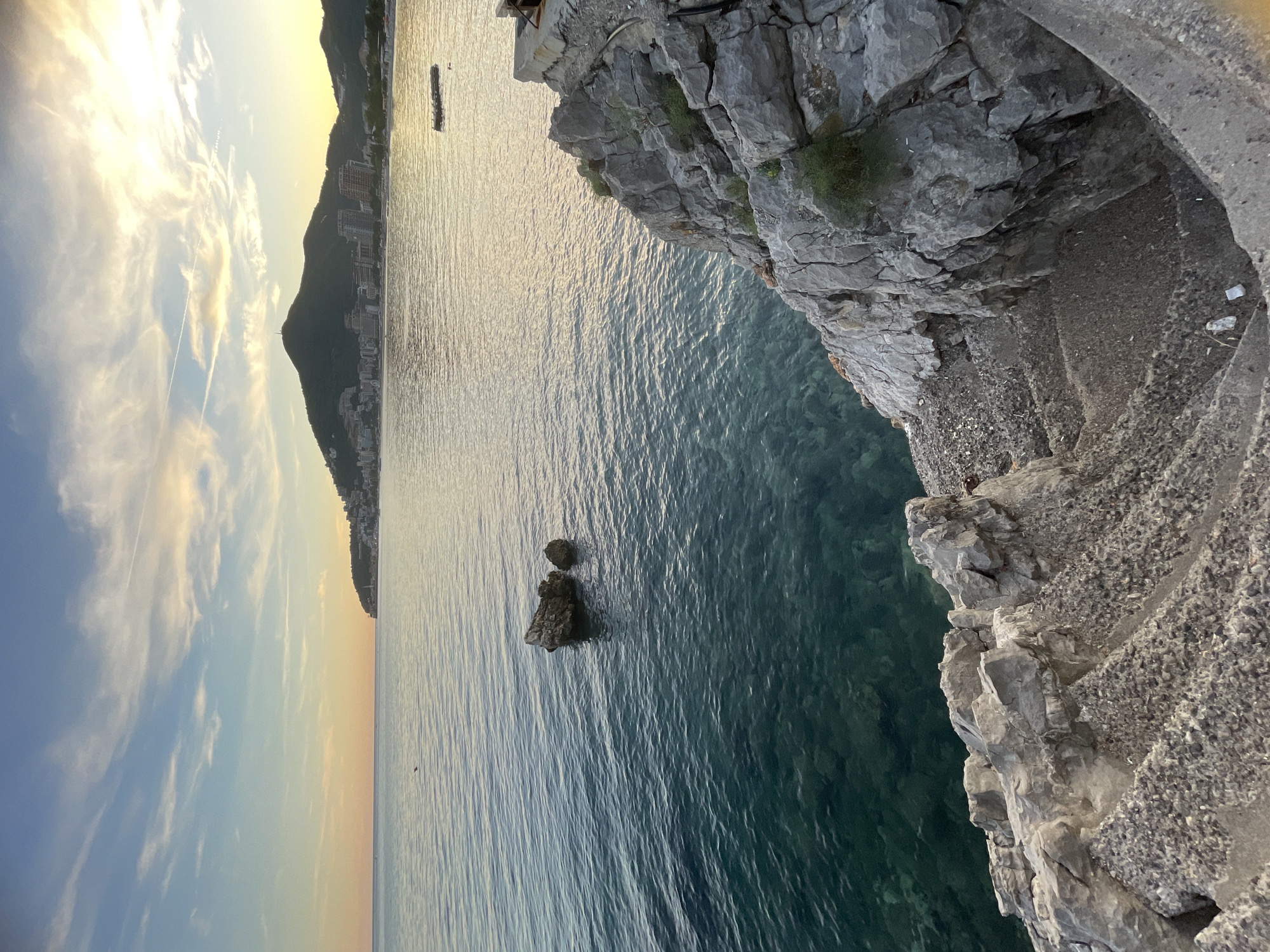 This image depicts a serene coastal scene at sunset in Budva, Montenegro. In the foreground, rugged, light-grey rocks form a steep embankment, with weathered stone steps leading down towards the water. Sparse green vegetation clings to the rocky surface.
The clear, turquoise water of the Adriatic Sea fills the midground, revealing the rocky seabed beneath. Two medium-sized rocks emerge from the water, creating a natural landmark. In the distance, the coastline of Budva is visible, characterized by a densely populated hillside covered in buildings and a prominent, tree-covered mountain rising behind the city. The sky above transitions from a soft orange hue near the horizon to a pale blue, with wispy white clouds scattered across it. The sun, low in the sky, casts a warm, golden light across the water and the distant landscape, creating a tranquil and picturesque atmosphere. A small, dark object, possibly a buoy or a marker, is visible in the water near the distant shore. The overall impression is one of natural beauty and peaceful serenity at the end of the day.