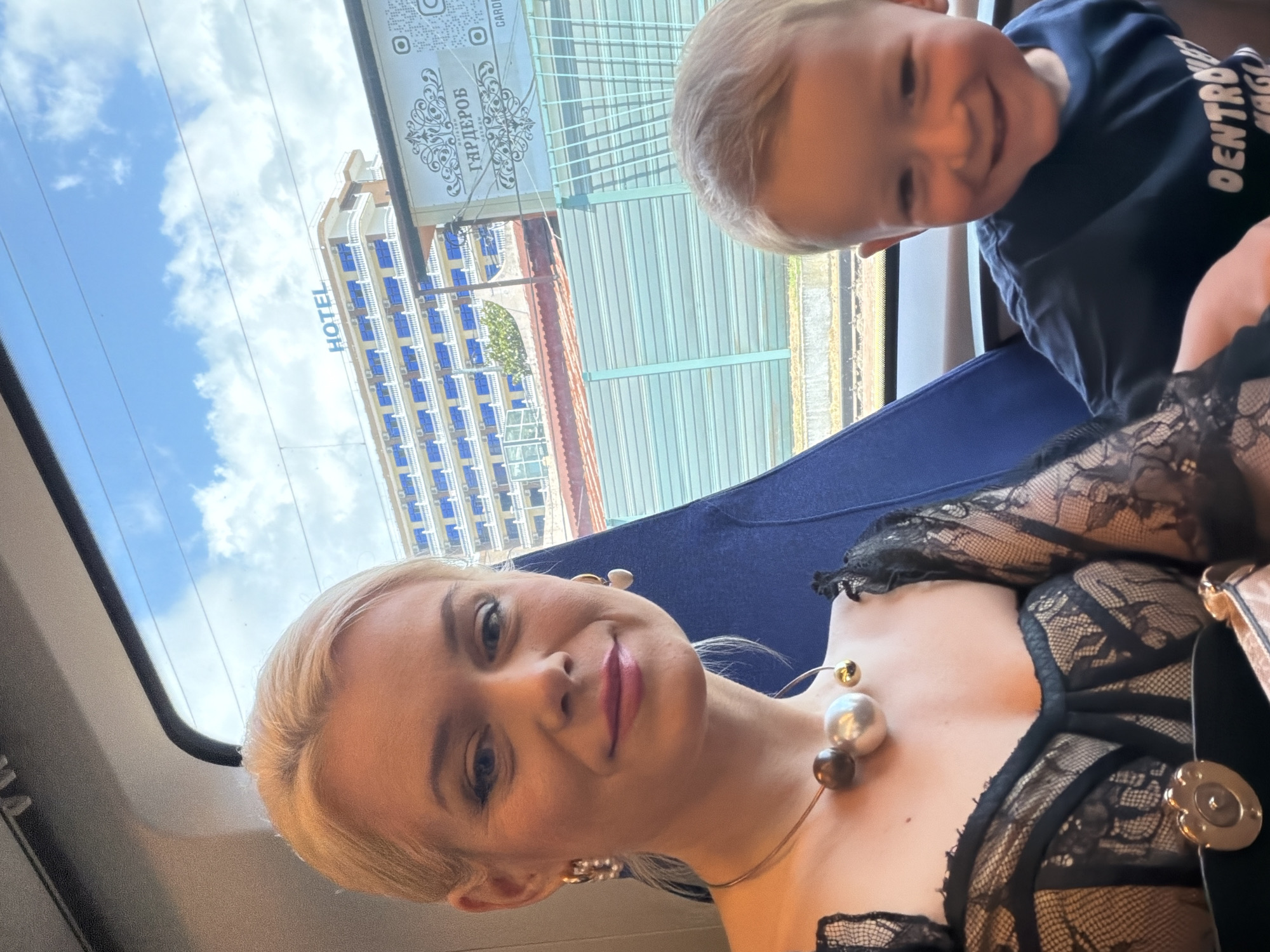 A young boy and a woman are seated together, likely on a train, as indicated by the view through the window. The boy, with blond hair, is looking towards the camera with a wide smile, wearing a dark blue shirt. The woman, with blonde hair, is also looking at the camera, smiling. She is wearing a black lace top and a necklace with large metallic beads.
Through the window, a tall building with many blue-windowed floors is visible, with the word "HOTEL" clearly printed on its side. Above this, a sign with Cyrillic text and decorative elements is partially visible. The sky is bright blue with white clouds, suggesting it is daytime and the weather is clear. The overall mood appears to be joyful and relaxed. The scene is set in Sochi, Russia.