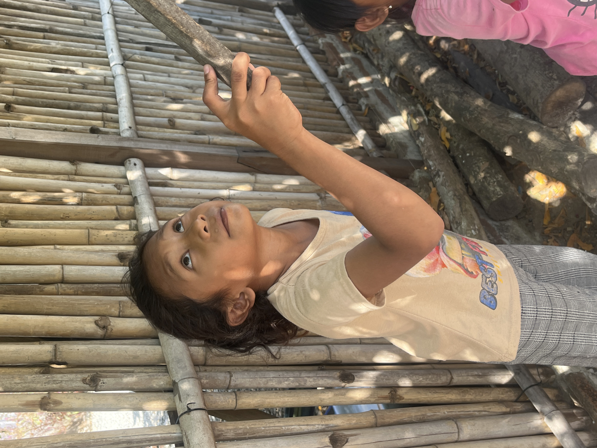 In Kertasari, Indonesia, a young girl with dark, curly hair tied back stands near a bamboo fence. She wears a light-colored t-shirt with a graphic print and grey checkered pants. Her gaze is directed upwards and to the right, her right hand extended to grasp a wooden stick. Sunlight casts shadows on her face and clothing. The background is dominated by the woven texture of the bamboo fence and a few scattered logs on the ground. To the right of the frame, a portion of another child wearing a pink shirt is visible.