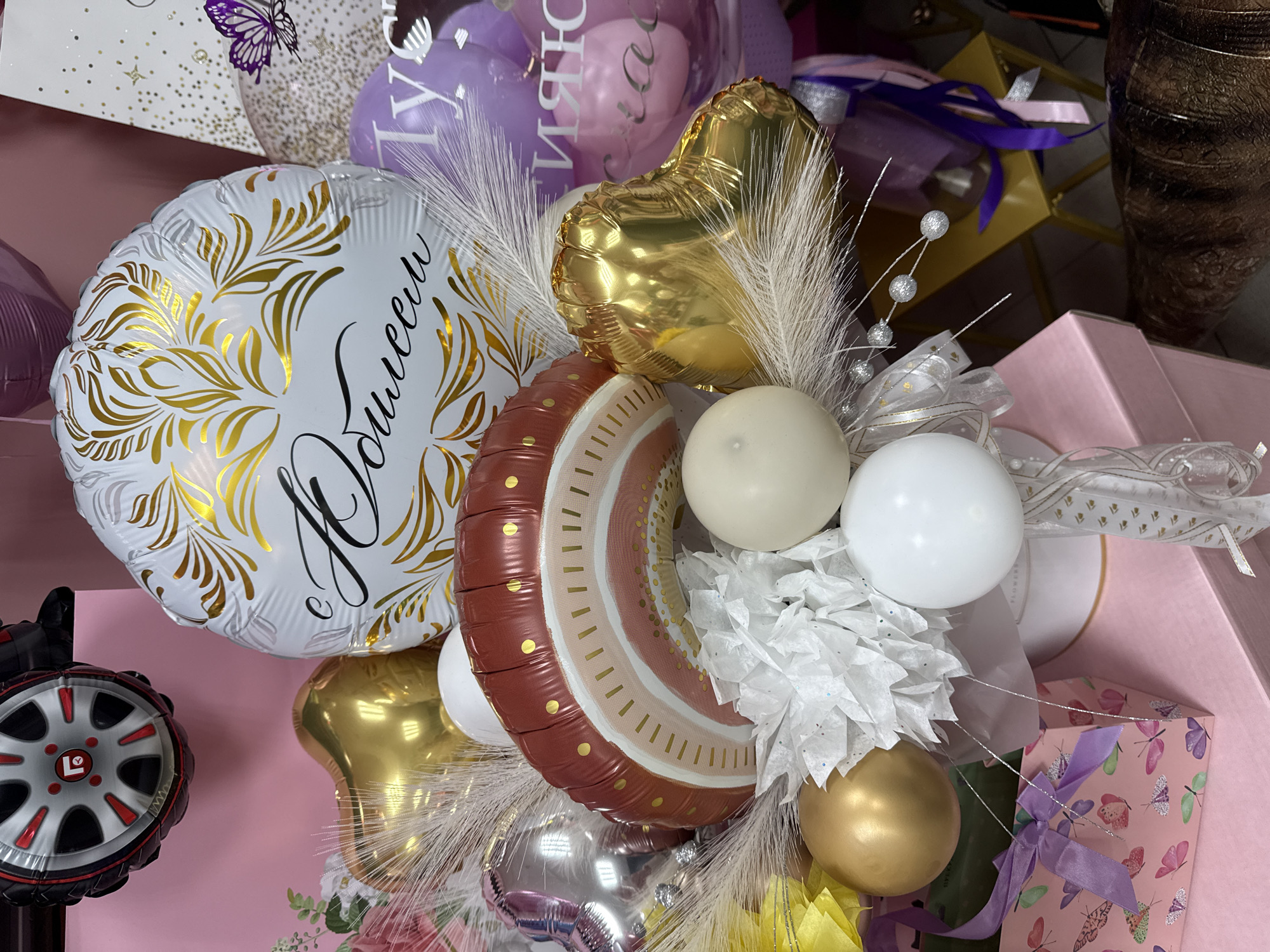 The image shows a close-up of a balloon bouquet with various types of balloons arranged together. A white round balloon with gold floral designs and black Cyrillic text is visible. Other balloons include a gold heart-shaped balloon, a brown and cream colored striped balloon with gold accents, and several smaller round balloons in gold, white, and cream colors. There are also white feathery decorations and white paper pom-poms interspersed among the balloons. The arrangement is placed among several pink gift boxes decorated with ribbons and butterfly patterns. The background is partially obscured, but hints of other balloons and possibly retail displays are visible, suggesting this scene might be in a gift shop. The location is specified as Unknown, Unknown.