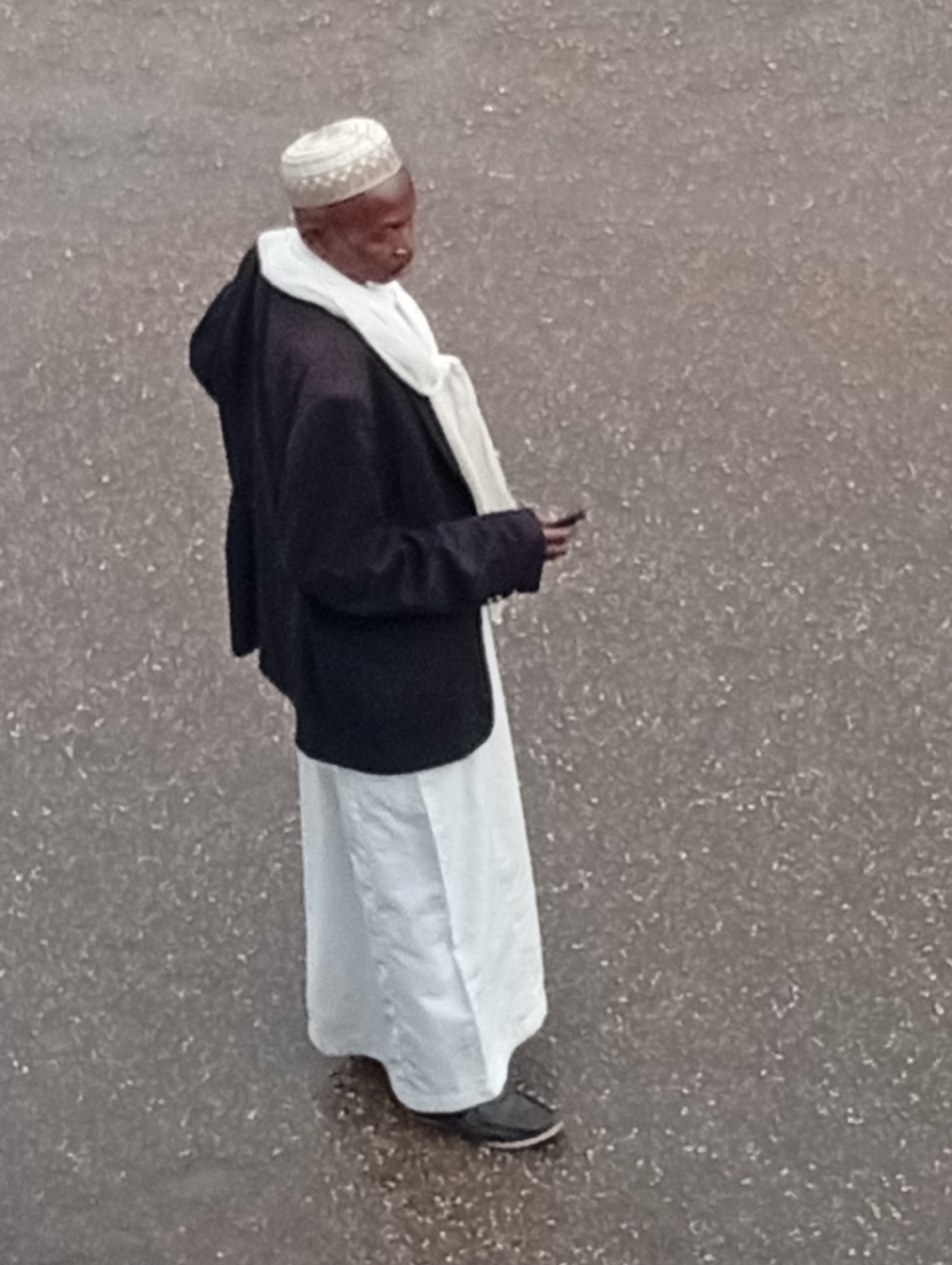 A person of African descent is standing outdoors in Bukuru, Nigeria. The person is wearing a white embroidered hat, a white scarf, a dark jacket, and a long white garment. The person is holding a mobile phone in their right hand and is looking down at it. The ground is covered with grey asphalt.