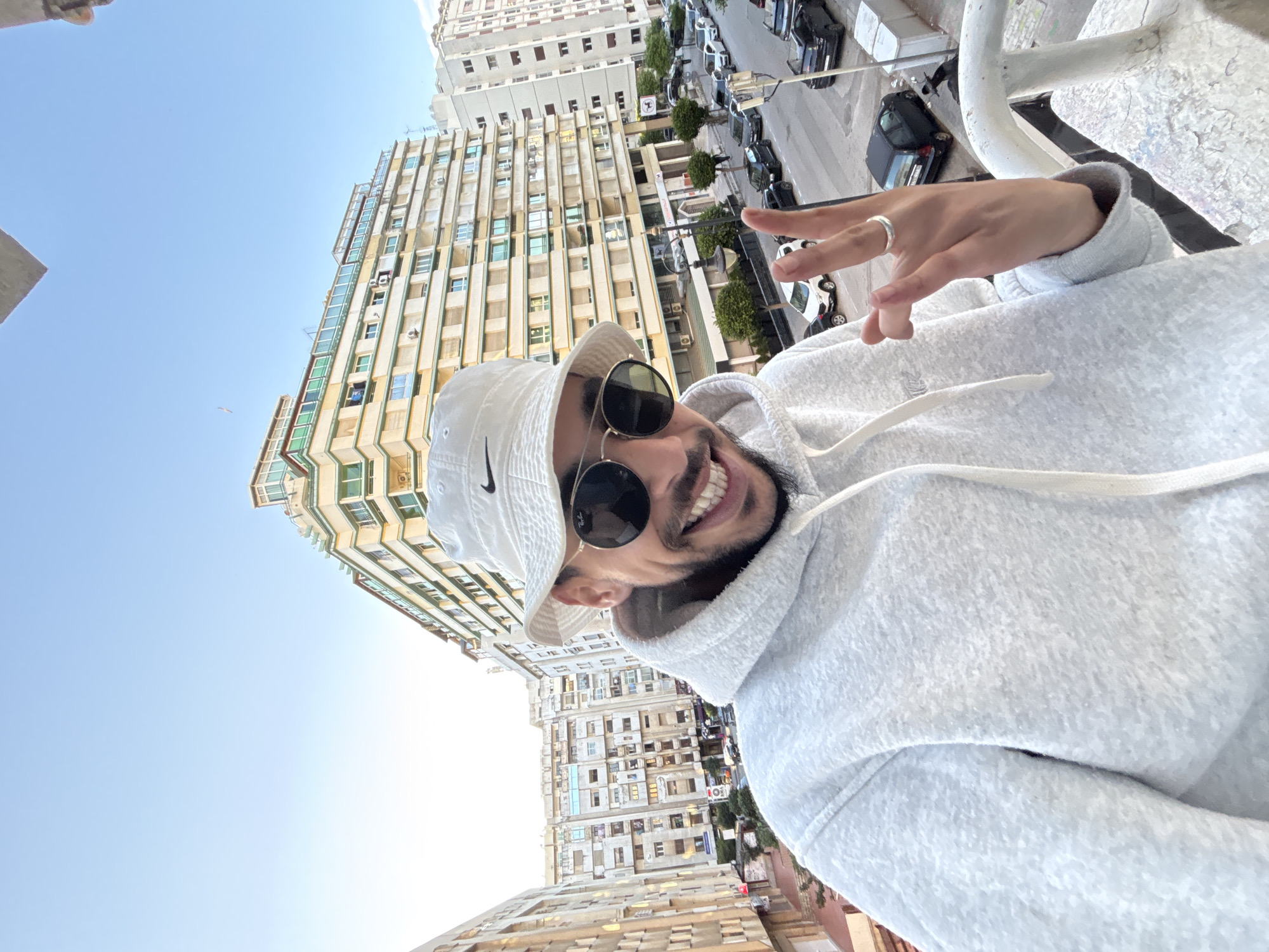 The image depicts a male subject taking a selfie from an elevated position, likely a balcony, overlooking a street and buildings in Tanger, Morocco. The subject is centrally framed, smiling broadly and showing teeth. He wears a white Nike bucket hat, dark circular sunglasses with a visible Ray-Ban logo, and a light grey textured hooded sweatshirt. His right hand is raised, making a "peace" gesture with the index and middle fingers, displaying a silver ring on his right ring finger.
The urban background under a clear, light blue sky features multiple multi-story buildings. To the left, an older, weathered residential building with numerous windows is visible. To the right, a more architecturally distinct building with light yellow facades, green-tinted windows, and a tiered upper structure dominates the skyline. Below, a street is visible, lined with parked vehicles including several dark-colored cars and a white SUV. Streetlights, small trees, and additional residential buildings are present further down the street. A weathered white metal railing of the balcony is partially visible in the lower right foreground.