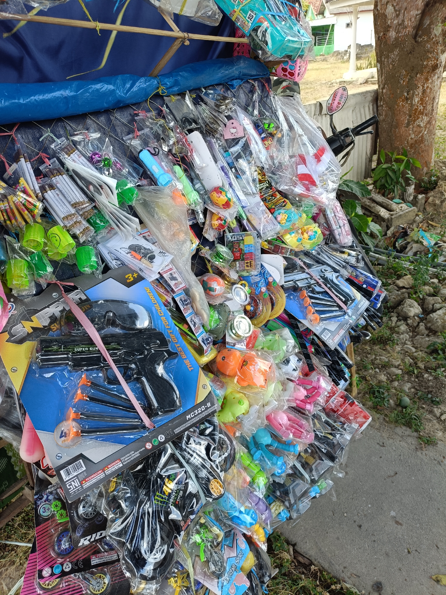 The image shows a street vendor's stall in Pordapor, Indonesia, overflowing with an assortment of packaged toys. The stall is set up outdoors, with a blue tarp providing shade and supported by bamboo poles. A variety of plastic toys are displayed, including toy guns, toy motorcycles, toy animals, small flashlights, and other novelty items. Many of the toys are still in their original plastic packaging, some with colorful graphics and warnings. The stall is positioned along a concrete walkway, with a dirt and gravel area visible to the right, dotted with green vegetation and a large tree. Part of a motorcycle is visible on the right side of the image, suggesting the stall might be set up near a road or parking area. The overall scene depicts a typical roadside market offering a wide selection of affordable toys.