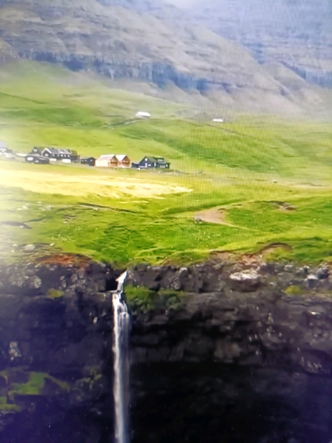 The image depicts a serene natural landscape with a waterfall in the foreground and a cluster of buildings nestled in a green, rolling hills in the background. The waterfall cascades down a dark, rocky cliff face, with vibrant green moss clinging to the rocks. Above the waterfall, the terrain opens up to a vast expanse of lush green fields, dotted with several small, uniformly designed houses. These houses have dark roofs and light-colored walls, suggesting a modern architectural style. In the far distance, a steep, gray mountain range forms the backdrop, its slopes textured with visible strata. The sky is overcast, contributing to a soft, diffused light that bathes the entire scene. The overall impression is one of a remote, idyllic settlement integrated harmoniously with its natural surroundings. There is no indication that this location is in Netanya, Israel.
