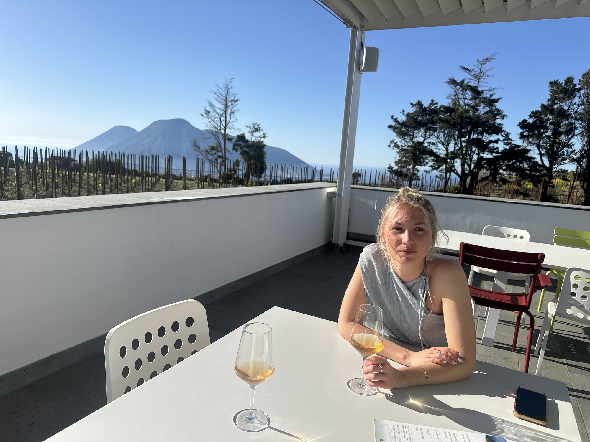 The image displays a woman seated at a white outdoor table on a sunny day, likely on a balcony or terrace with a panoramic view. She has blonde hair and is wearing a light blue top, holding a glass of what appears to be rosé or white wine. Another similar glass sits on the table in front of her, alongside a menu or paper and a smartphone.
The setting is an elevated outdoor area, possibly a restaurant or winery in Lipari, Italy, overlooking a vast body of water and a distinctive mountainous landscape featuring two prominent peaks under a clear, bright blue sky. A fence-like structure, reminiscent of a vineyard, borders the foreground, with various trees dotting the scene. The terrace itself has white walls and a light-colored overhead structure, with a speaker mounted on a pillar. Other empty chairs in white, dark red, and lime green are visible further back on the terrace.
The clear sky and sharp shadows suggest a bright morning or afternoon. The woman has a calm and pleasant expression, looking directly towards the camera.
Visible text on the menu includes items such as "BANCO", "ROSA CACLINO", "MALIRSIA DELLE LIAR", "ANALOCHLIC DRINK", "BREAD BUTTER AND ANCHO", and "COLD CUTS AND CHEESES PLA".