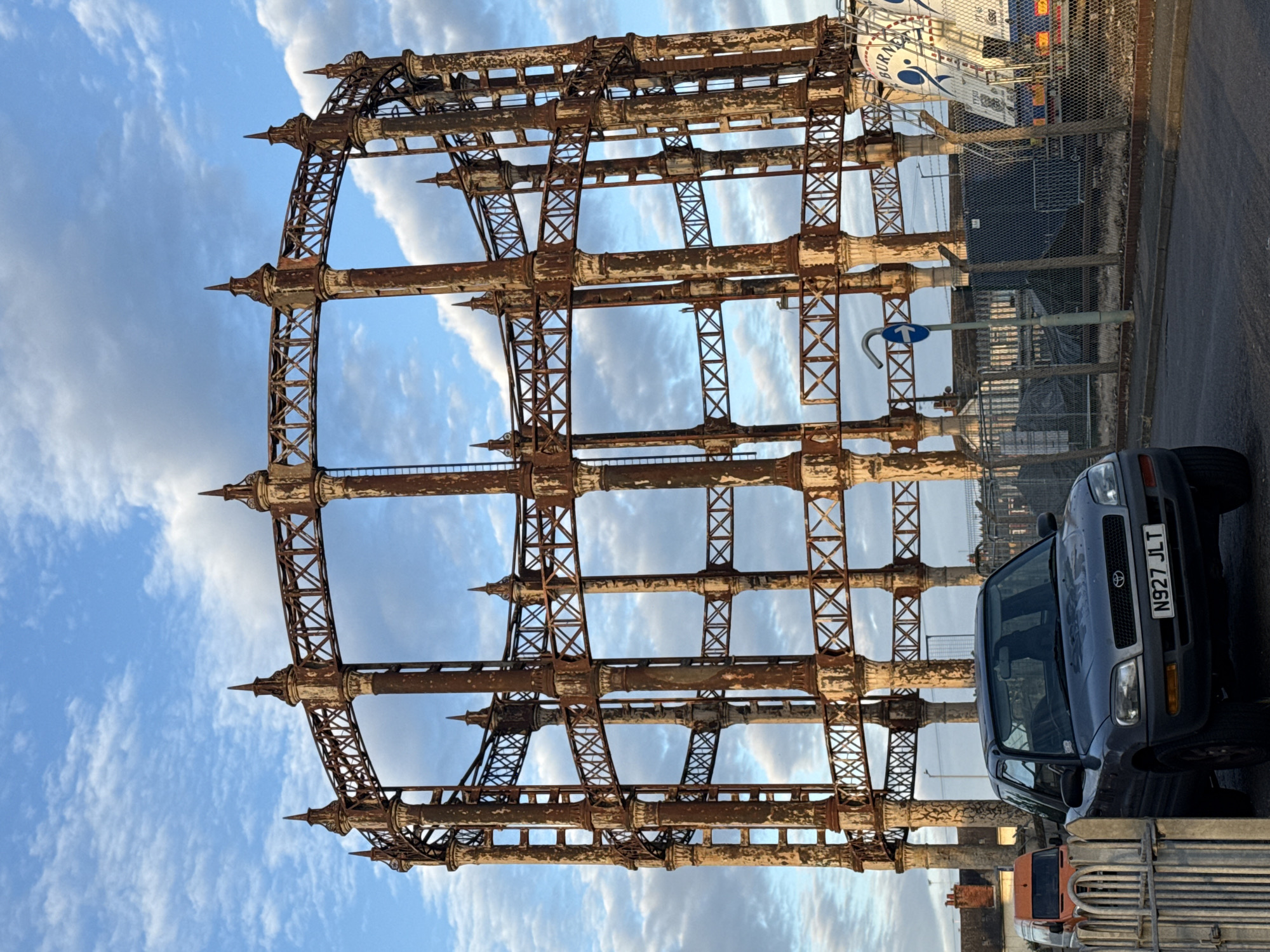 The image shows the rusty skeletal structure of a former gas holder in Great Yarmouth, United Kingdom, set against a blue sky with scattered clouds. The frame is constructed of vertical and horizontal pillars connected by cross-bracing. A gray Toyota RAV4 with the license plate N927 JLT is parked to the right of the structure. The background includes a chain-link fence, a portion of a large white truck, and a blue and white traffic sign.