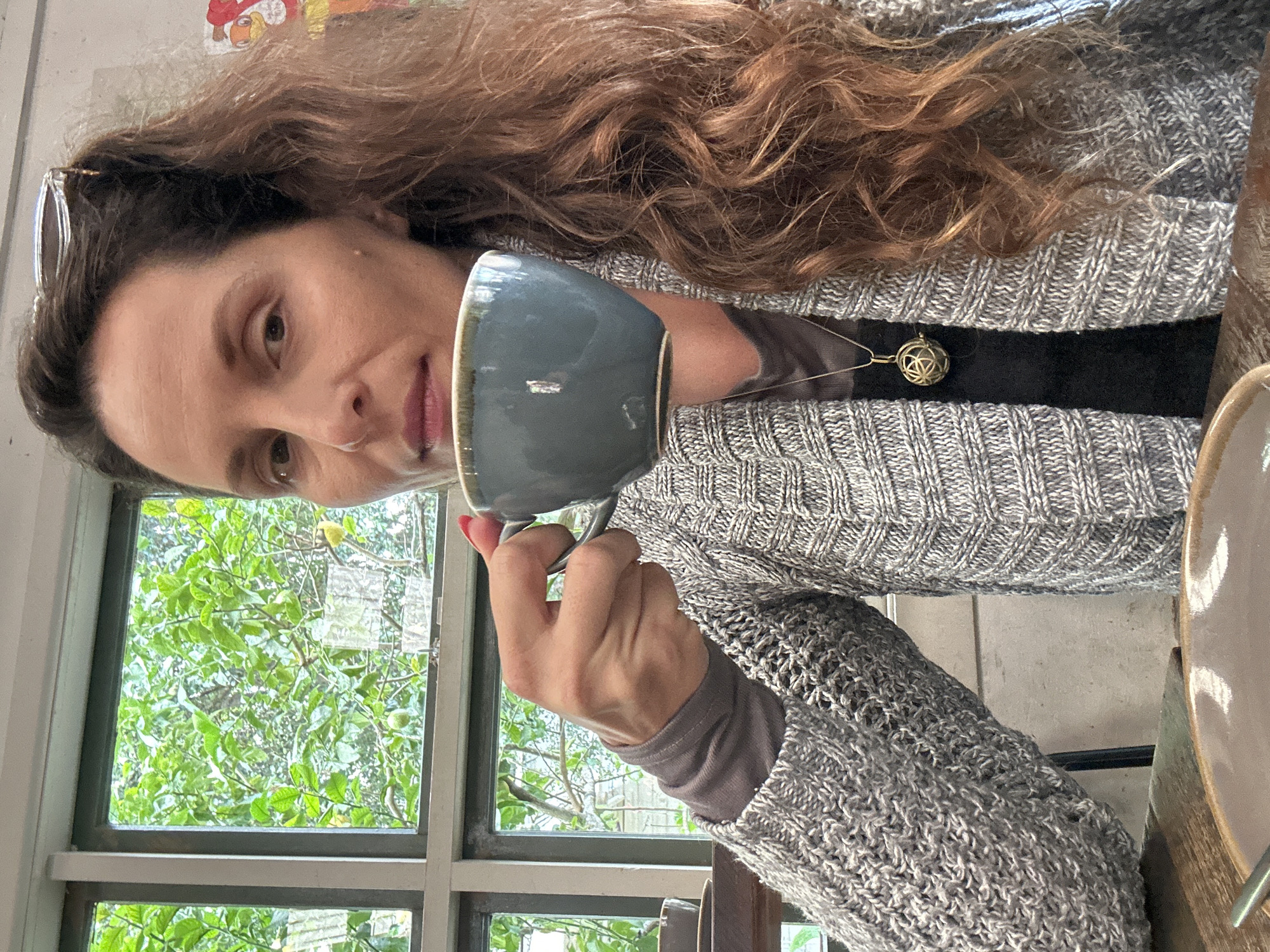 The image shows a woman holding a grey cup of coffee in her right hand. She is wearing a grey knitted cardigan over a dark top and a pendant necklace. She is sitting at a wooden table in front of a window with greenery visible outside. The scene appears to be set in a cafe. The woman has long, wavy brown hair and is looking towards the camera. The cup is ceramic with a blue-grey glaze and a brown rim. A plate with a light brown ceramic bowl is also visible on the table. The background shows a window with multiple panes, revealing lush green trees and foliage. A hint of what might be a colourful artwork or poster is visible on the wall to the right. The lighting suggests it is daytime. There is no specific indication of the city of Bellingen, Australia, within the image itself.