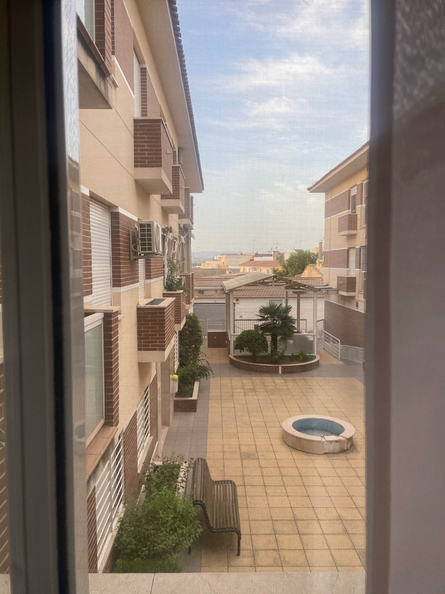 This image depicts a view of a courtyard between buildings, likely an apartment complex, in Atarfe, Spain. The foreground is dominated by the edges of a window frame, with a visible screen distorting the view.
The courtyard itself is paved with light-colored square tiles and features a small, oval-shaped water feature, possibly a decorative pond or a small, shallow pool, in the center. To the left of the water feature, there is a small palm tree surrounded by a low, circular brick border and some low-lying bushes.
On the left side of the courtyard is the facade of a multi-story building, characterized by light beige walls with brick accents around windows and balconies. Several air conditioning units are mounted on this building. White roller shutters are visible on some of the windows.
On the right side of the courtyard, another building with a similar architectural style is visible, featuring brick detailing and balconies. Further in the background, other buildings with red-tiled roofs are seen, extending into the horizon under a partly cloudy sky. A wooden bench is positioned in the foreground on the right, adjacent to a patch of dense green bushes. A thin, tall fence or railing runs along the right edge of the courtyard, leading towards the background. The overall scene suggests a residential area, possibly with shared outdoor space.