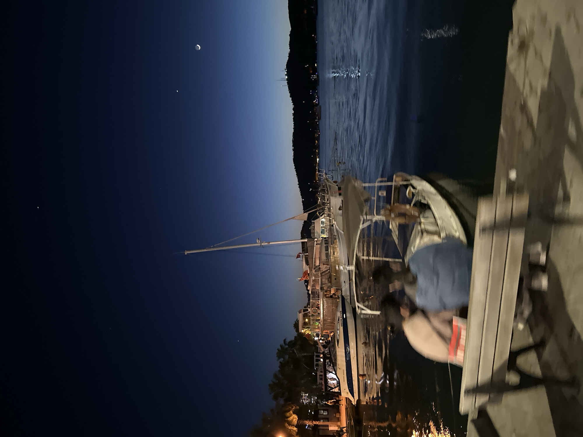 The image depicts a nighttime scene in Fethiye, Turkey. A sailboat is moored along the harbor or marina with other boats, positioned against the backdrop of a dark blue sky punctuated by a crescent moon and a few visible stars. The opposite side of the harbor has a dimly lit skyline. On the dock, a bench is visible, partially occupied by indistinct figures who appear to be observing the scene. Lights from the boats and shoreline buildings reflect in the water, creating a series of blurred golden and white spots. The photograph is taken at an angle, making the scene appear sideways.