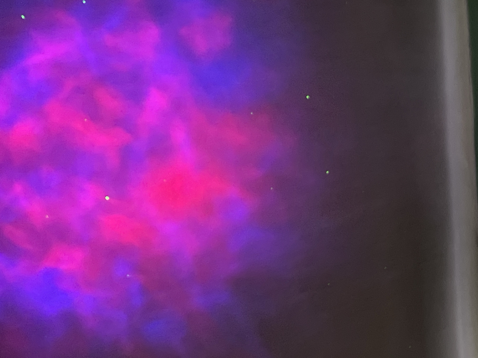 The image shows a blurred, nebulous pattern of pink and purple hues. There are also a few scattered, small green points of light. Part of a solid, light-colored vertical surface is visible on the right edge of the image. The location is not apparent, and cannot be determined from the visual content. No people, subjects, objects, actions, or interactions are visible.