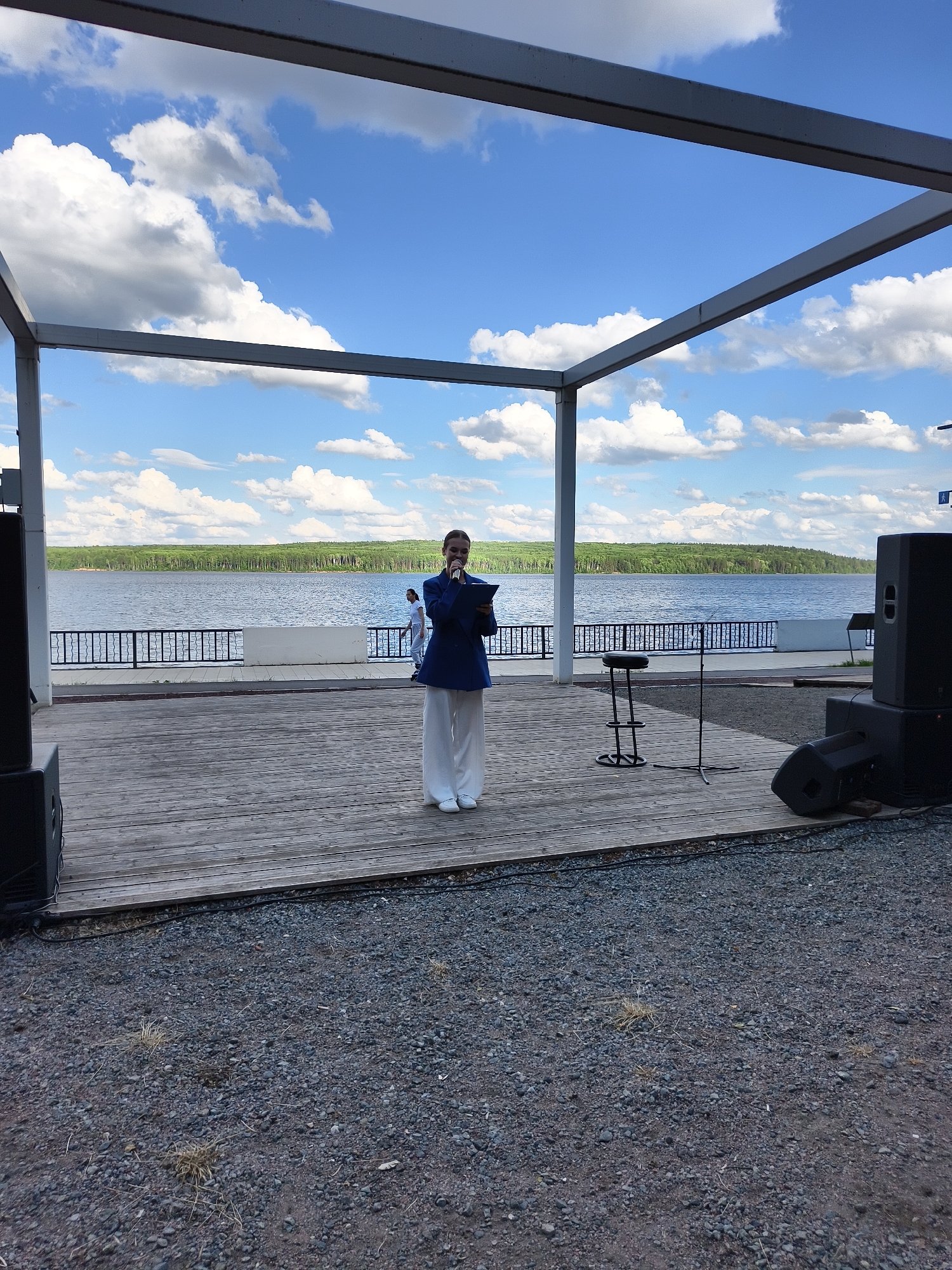 A person, appearing to be female and dressed in a dark blue blazer and white wide-leg pants, stands on a wooden stage holding a tablet. She is speaking into a microphone positioned on a stand. Behind her, a body of water is visible, with a line of trees along the far shore. The sky is blue with scattered white clouds. The stage area has two large speakers on either side, and a stool and another microphone stand are also present. The foreground is covered in gravel.