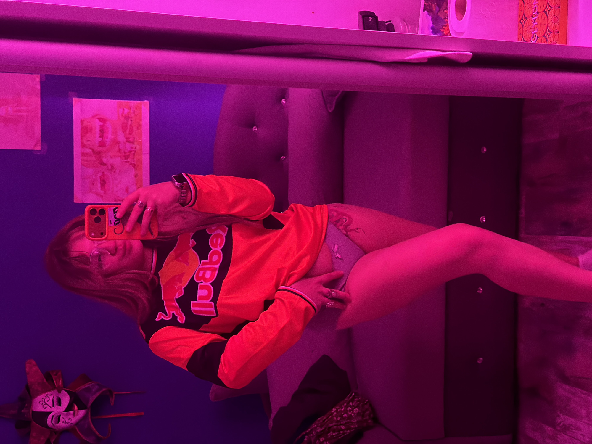 A person is visible in a mirror selfie inside a room. The room appears to be illuminated with purple and pink lighting. The person is holding a phone, and wearing an orange and black long-sleeved shirt with the word "Redbull" on the front. They are also wearing grey bottoms. Their hair is blonde and they have glasses on. A decorative Venetian mask is visible on the wall in the lower left corner. There are also pictures hanging on the blue wall behind them. A cushioned headboard is behind the person. The location is not discernible.