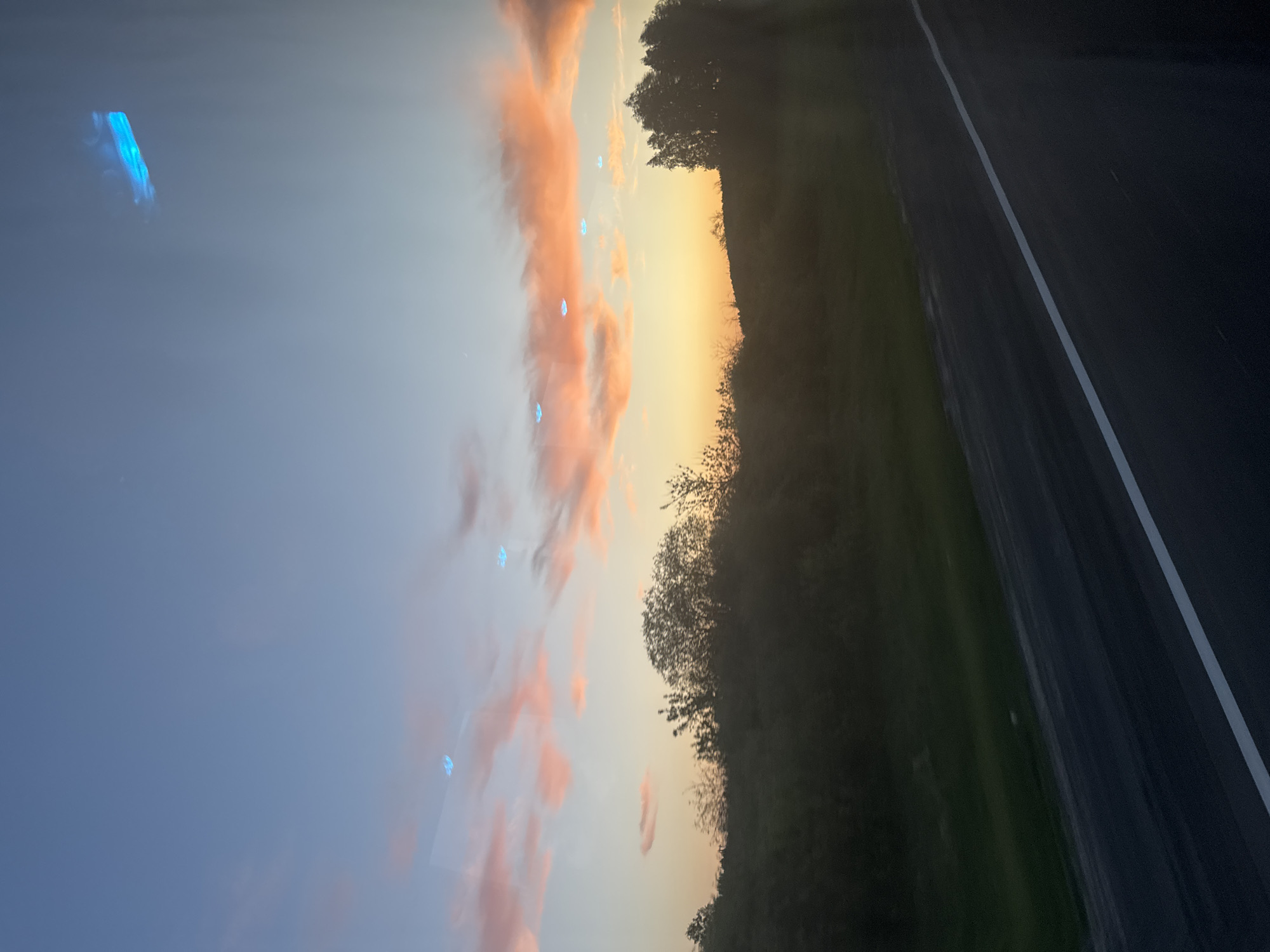 The image depicts a roadside view at sunset or sunrise. The sky is predominantly blue with pink-tinged clouds. The horizon displays a transition from yellow to orange hues. A dark green area with trees is visible along the roadside. The road surface is black with a white lane marking. The image was likely taken from inside a moving vehicle. The presence of a road and trees suggests a location outside of a densely populated city. Without further information, it is not possible to confirm that the image was taken in Федькино, Russia.