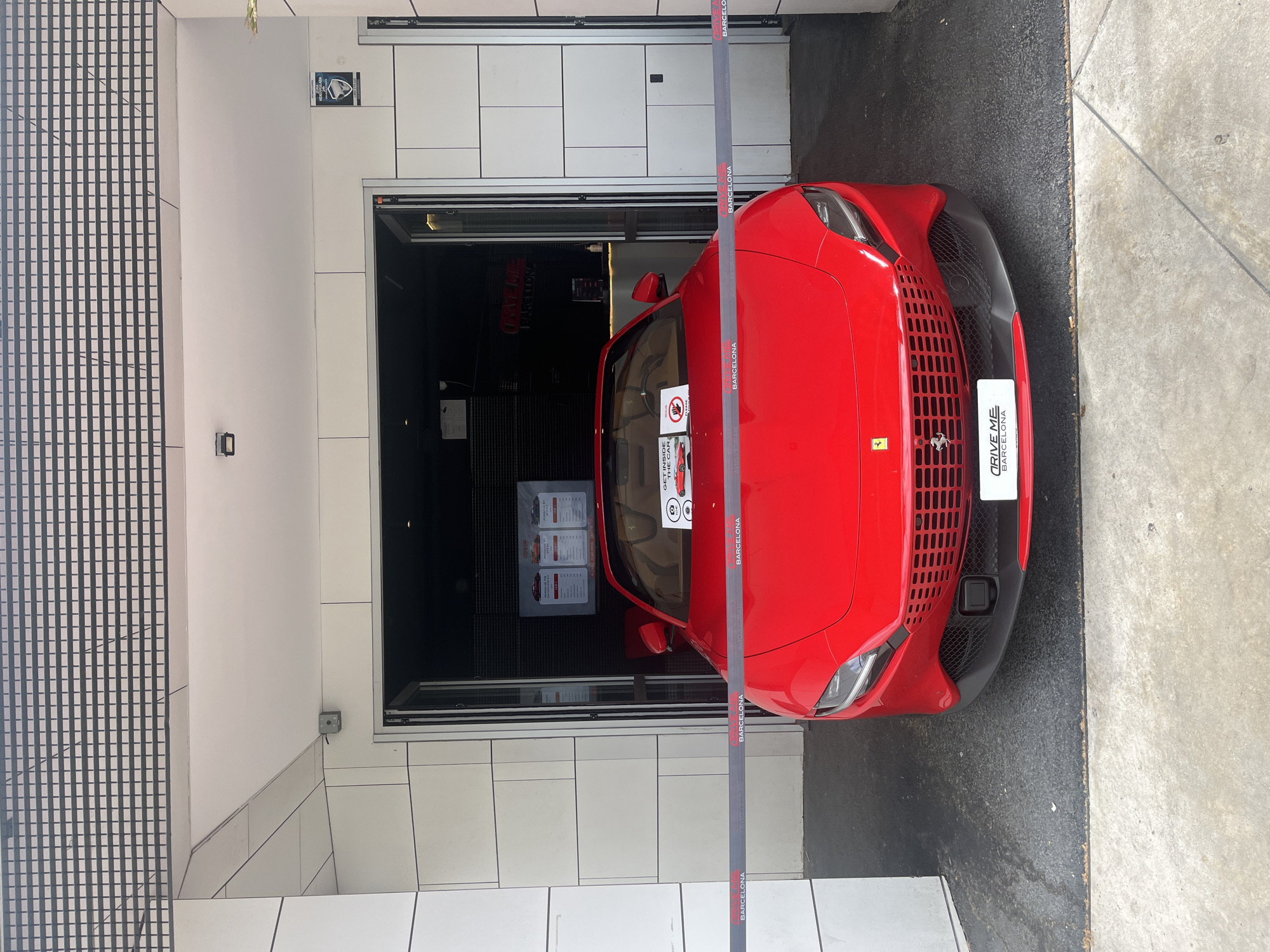 A vibrant red sports car, identifiable as a Ferrari by its emblem on the grille, is prominently displayed at the entrance of a garage or showroom. The car is positioned with its front partially extended outside the building. A grey ribbon, repeatedly featuring "BARCELONA" in white text, stretches across the front bumper and along the side of the vehicle, serving as a barrier or promotional element.
Affixed to the car's front bumper is a white sign that clearly states "DRIVE ME BARCELONA." On the driver's side window, a smaller white sign is visible, partially reading "GET INSIDE THE CAR" and depicting a 'no touching' symbol, suggesting it's part of an interactive display for a driving experience.
Inside the dark garage, several posters with unreadable text (likely outlining pricing or package details) are visible behind the car. The exterior of the building features tiled walls in white and grey, with a distinct grid pattern on the left side. A small security camera is mounted on the wall to the left of the garage entrance. The ground transitions from dark asphalt directly in front of the garage to lighter concrete pavement further out.
The scene is set during the daytime, indicated by the clear and bright lighting, suggesting pleasant weather in Barcelona, Spain. The overall context points to a luxury car rental or driving experience service.