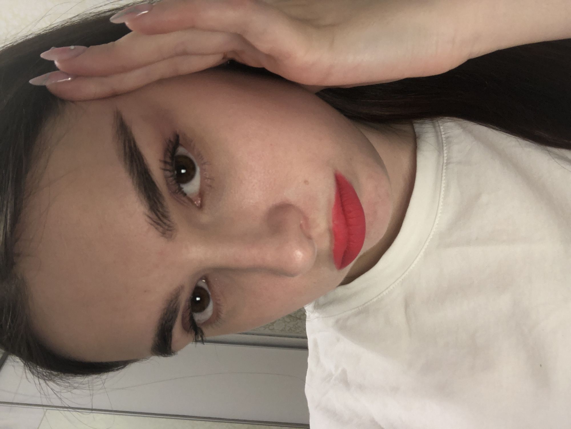 A close-up portrait captures a young woman's face, oriented with a slight tilt, as she looks directly towards the viewer. She has fair skin, dark brown eyes with visible mascara on her eyelashes, and prominent, well-defined dark eyebrows. Her lips are painted with a vibrant, matte red lipstick. Dark hair is visible framing her face and flowing over her shoulder.
Her right hand is gently placed across her forehead, with her long, almond-shaped fingernails resting near her hairline. The nails are painted in a light, possibly nude or translucent pink shade, with subtle glitter or decorative accents. She is wearing a simple white t-shirt, visible around her neck and upper chest.
The background is softly blurred and indistinct, featuring light-toned surfaces that suggest an indoor setting. The lighting appears soft and even, highlighting her facial features. There are no specific objects or identifiable text visible in the background. The image conveys a neutral or contemplative expression, focusing on her striking makeup and the detail of her manicured hand. The image itself provides no explicit geographical cues beyond the provided context of Noviy, Russia.