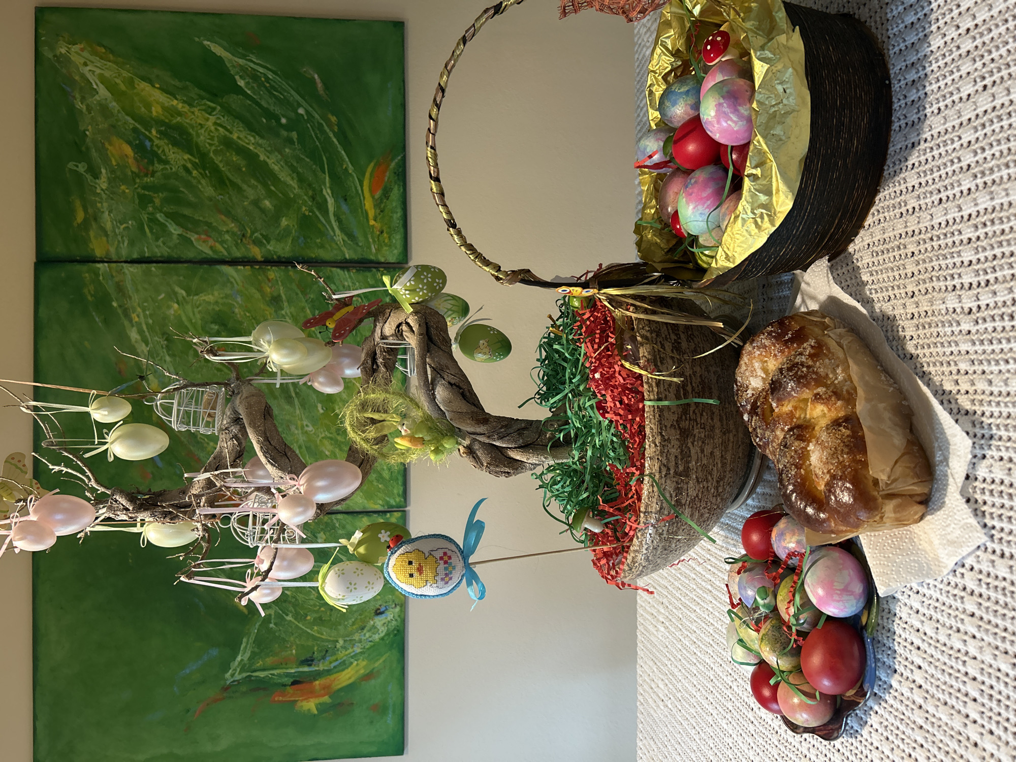 The image shows an Easter-themed still life arrangement on a white textured surface. In the background, there is a two-panel abstract painting with green, yellow, and white tones.
The still life features an Easter egg tree with hanging pastel-colored eggs and small birdcage decorations. A woven basket filled with brightly colored Easter eggs in golden foil is positioned to the right. Beneath it, there is a braided sweet bread wrapped in paper. Another small bowl of painted eggs with green ribbons is present.
Red and green shredded paper decorations are scattered around the arrangement. The location in the city of Unknown, Unknown is not determinable.