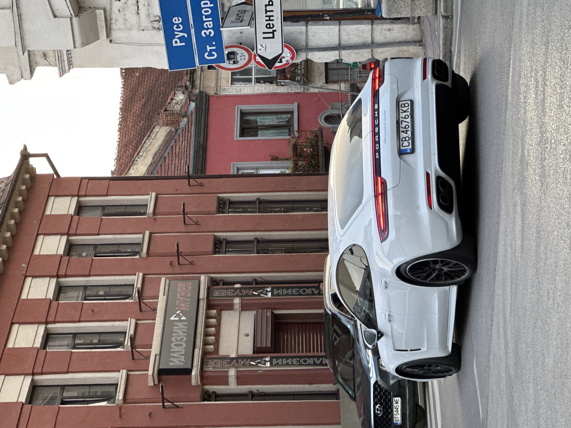 The image shows a white Porsche Panamera driving on a street in Veliko Tarnovo, Bulgaria. The car's license plate is visible and reads "CB 4676 KB". In the background, there is a red building with Cyrillic signage that appears to be an illusion museum. There are also street signs visible, one pointing to "Ruse" and "St. Zagora", and another to "Center" with a directional arrow. The street appears to be an intersection. The lighting suggests it is daytime, possibly late afternoon or early morning due to the soft light.