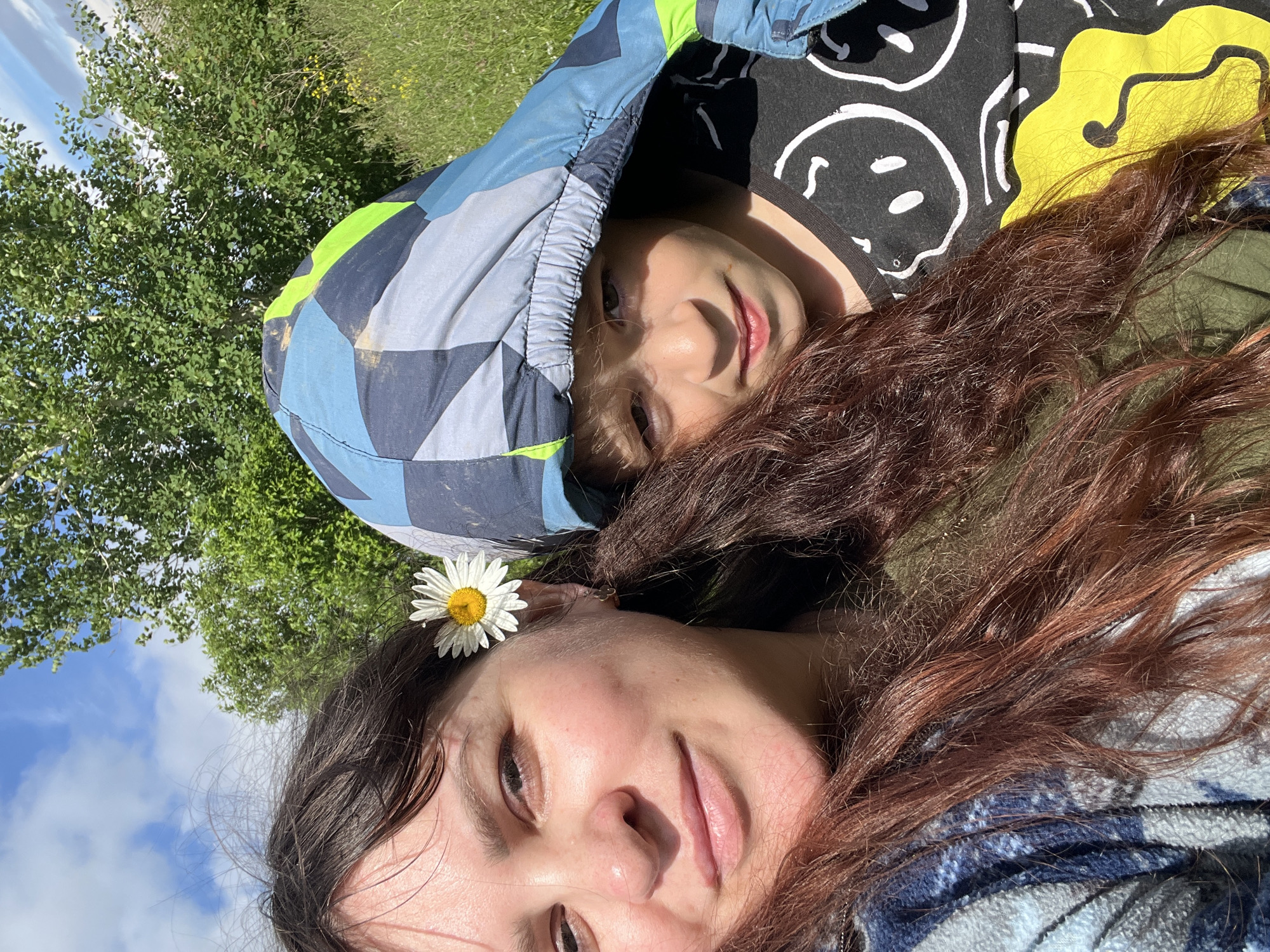 A selfie is taken by a woman with dark hair, a daisy in her hair, and a blue and gray checkered jacket. Next to her is a child wearing a patterned hooded jacket and a black t-shirt with smiley faces. They are outdoors in a grassy area with trees and a blue sky with clouds. The location is specified as Panteleevka, Russia.