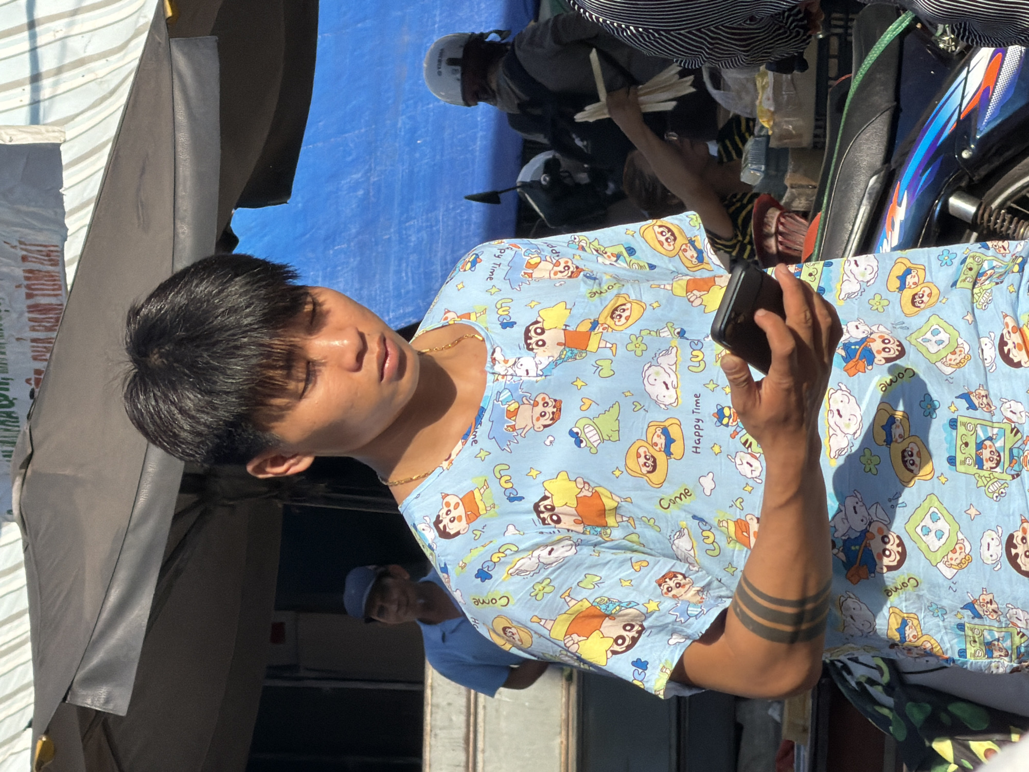 A young man with black hair and tattoos on his left arm stands in the foreground, holding a smartphone. He is wearing a light blue t-shirt with a cartoon print featuring characters like Shin-chan. The background is blurred, but suggests an outdoor market scene in Nha Trang, Vietnam, with awnings, tarps, and other people visible. One person in the background, wearing a blue shirt and cap, appears to be a vendor. Another person, wearing a helmet and backpack, is seen near a motorcycle. The overall lighting indicates it is daytime.