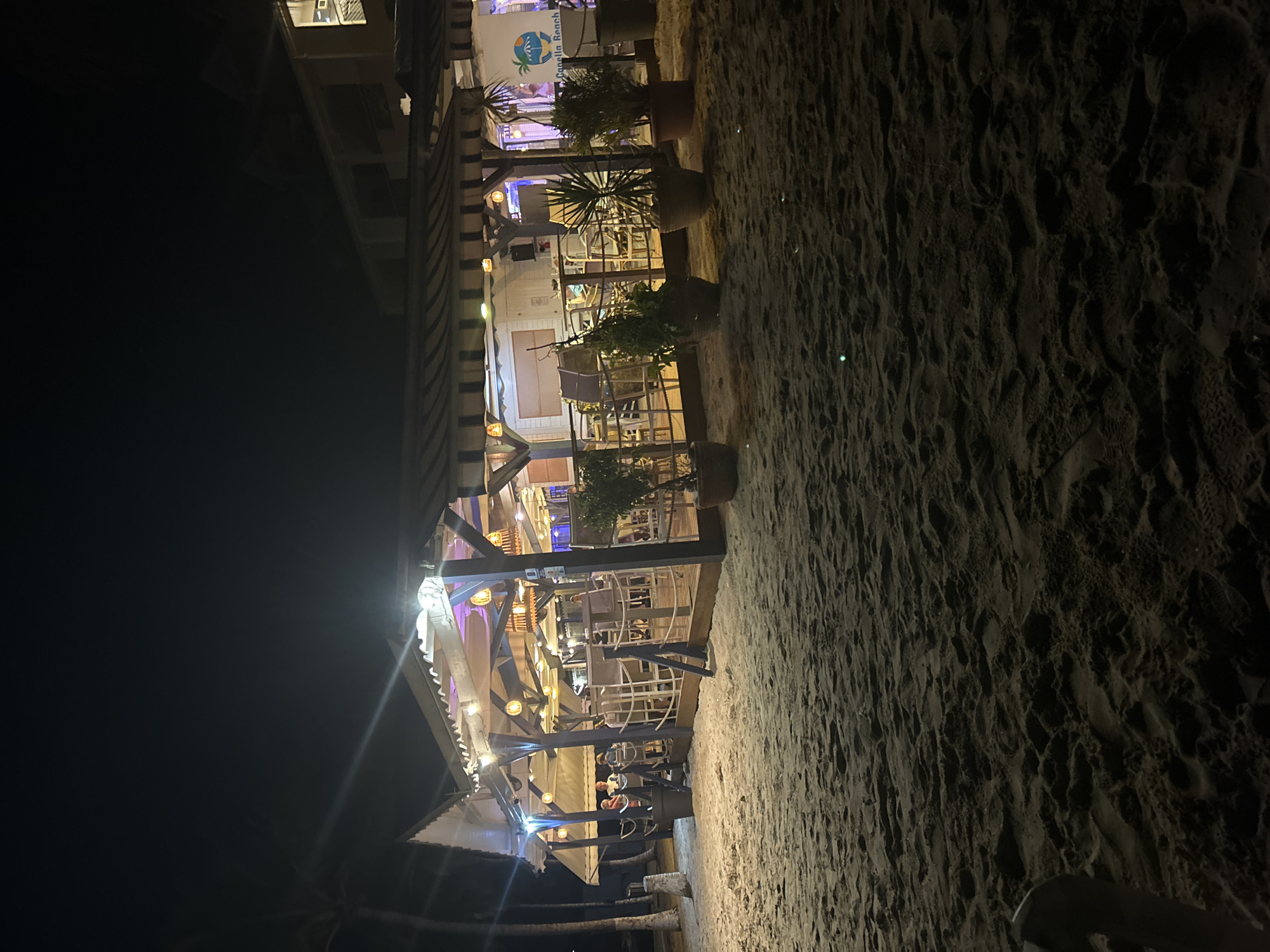 A nighttime view shows a beachside restaurant in Le Gosier, France, with lights illuminating the open-air structure. The sand of the beach forms the foreground, leading up to the building, which houses multiple tables and chairs, some occupied by patrons. Potted plants are situated along the edge of the restaurant, and a sign reading "Canella Beach" is visible. The night sky fills the upper left quadrant of the scene.