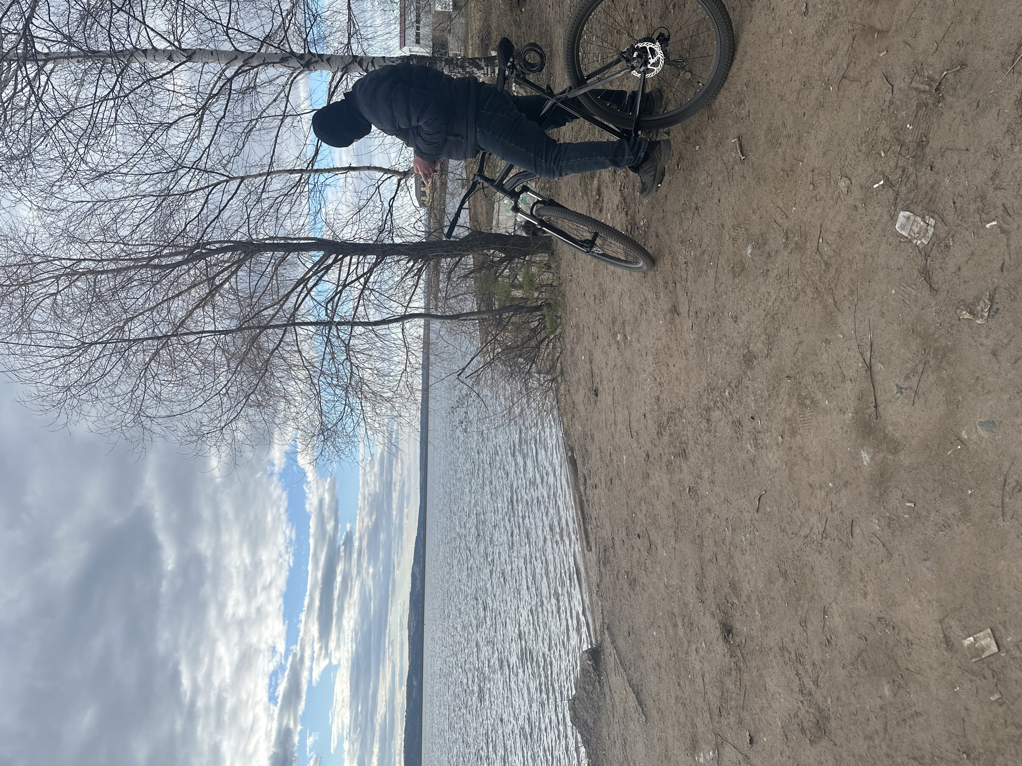 A man wearing a black jacket, black jeans, and a black hat stands beside a black bicycle on a dirt path. Behind him, a lake stretches into the distance under a cloudy sky. Bare trees are visible on the path and along the lakeshore. The location is likely near a body of water in Chaikovsky, Russia. The man is interacting with his phone.