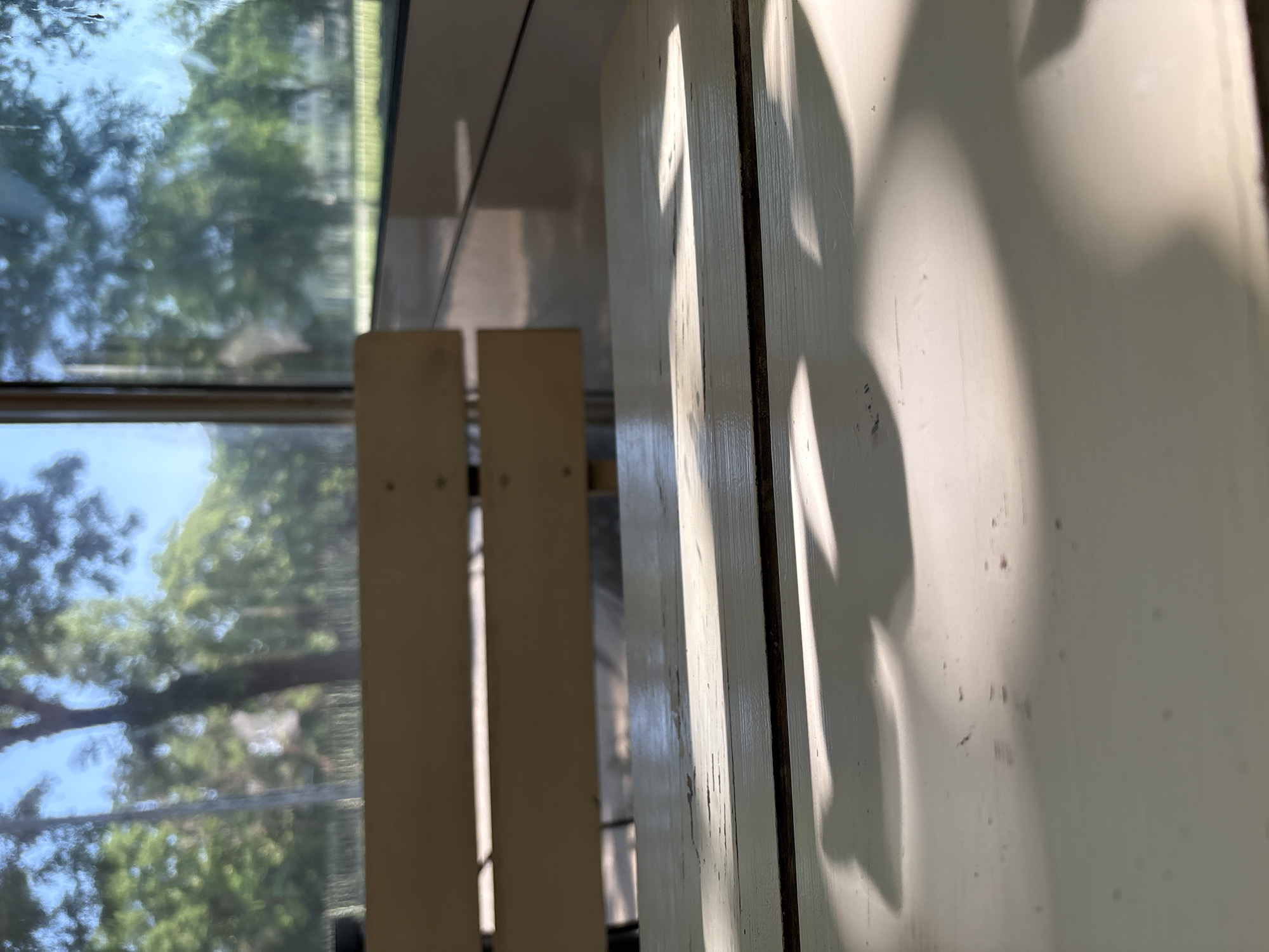 The image captures a close-up view of a weathered, light-colored wooden table, likely outdoors. The table's surface shows signs of wear and tear, with visible cracks and peeling paint. Sunlight casts dappled shadows across the table, suggesting it is daytime and there is a light source filtering through an overhead cover or trees. In the background, out of focus, a wooden bench is visible. Beyond the bench, a blurred scene of greenery and trees under a bright sky can be observed, hinting at a park or garden setting. There are no people or specific landmarks to identify the exact location within Plovdiv, Bulgaria. The overall impression is one of a simple, relaxed outdoor seating area.