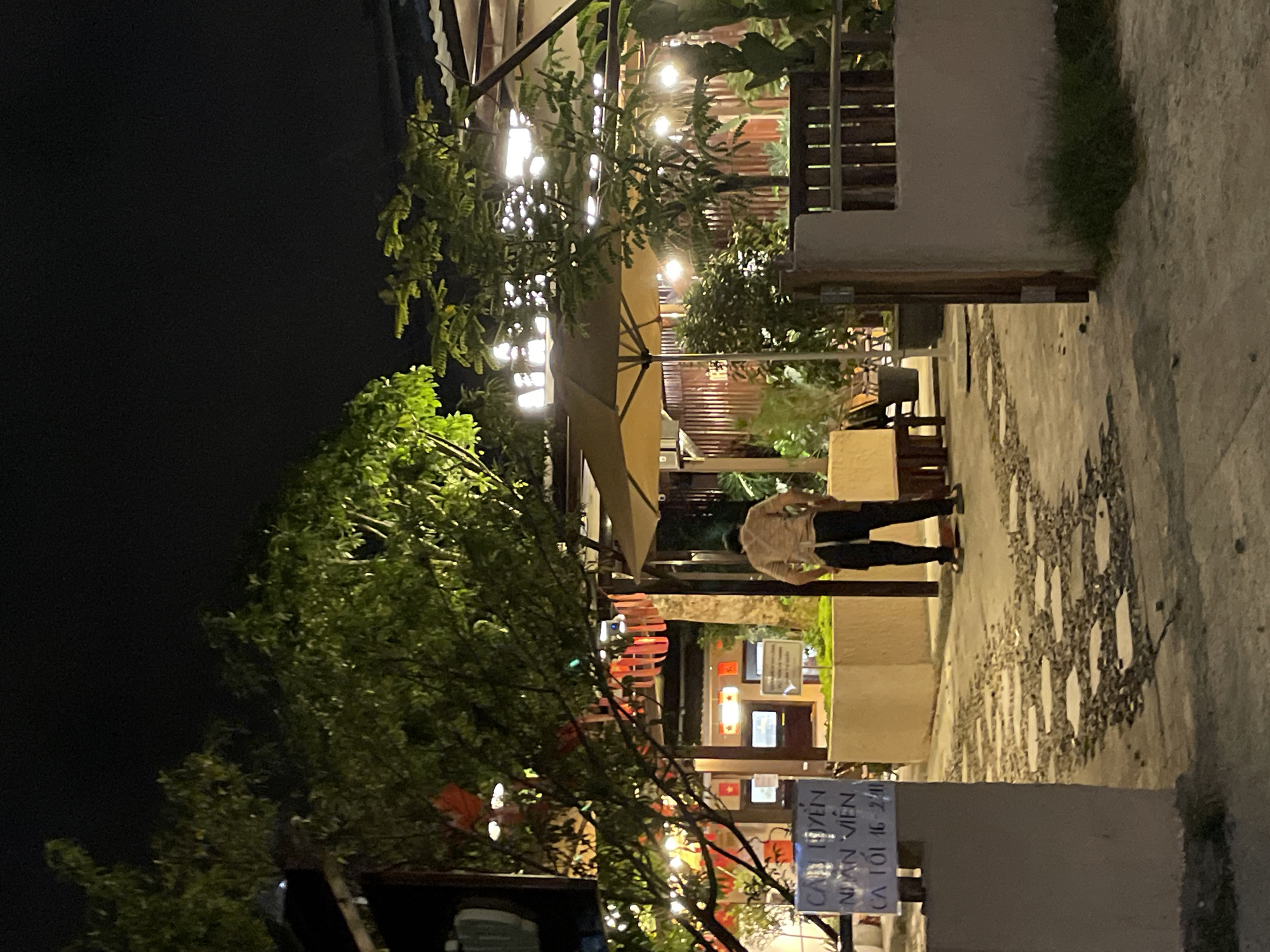 The image depicts an outdoor seating area of a establishment at night, likely in Lâm Viên - Đà Lạt, Vietnam, as indicated by the text on the sign. A person, presumably an employee, wearing a light-colored shirt and dark apron, stands in the center of the scene with their back to the camera. They appear to be tending to something near a planter box. The area is lit by string lights and ambient illumination from the building in the background, casting a warm glow on the stone-paved ground and surrounding vegetation. Several tables and chairs are visible, suggesting a dining or relaxation space. Decorative red lanterns hang from a tree, adding to the festive atmosphere. A sign with Vietnamese text is prominently displayed on the left. The environment appears to be a courtyard or garden setting, with trees and fencing contributing to the ambiance. The overall impression is one of a moderately busy, dimly lit, outdoor venue.