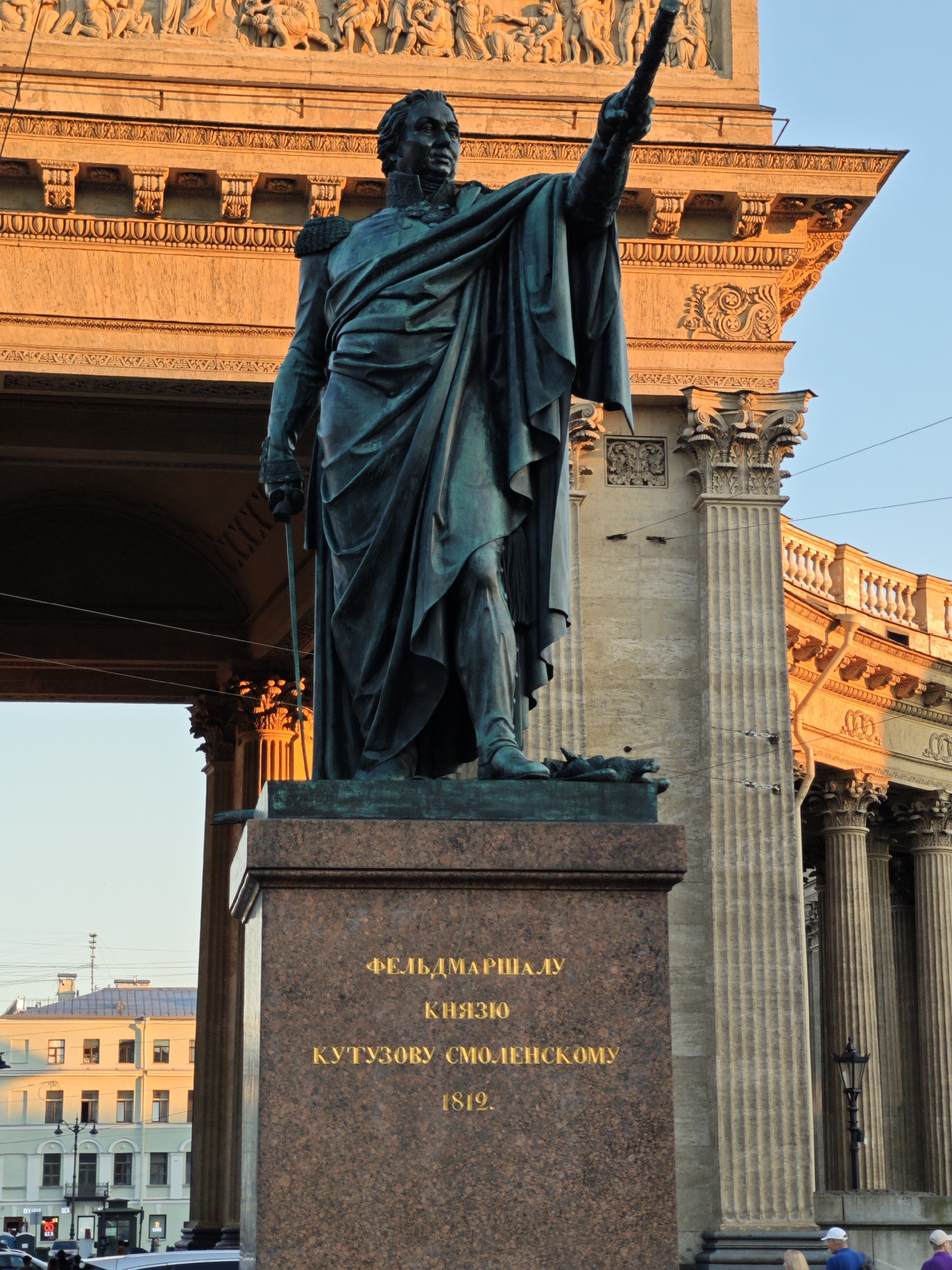 The image displays a low-angle, full-length view of the bronze monument to Field Marshal Mikhail Kutuzov, situated in front of the Kazan Cathedral in Saint Petersburg, Russia. The statue depicts a male figure in military attire with a long, draped cloak, holding a sword in his left hand and pointing forward with his raised right arm. The bronze surface exhibits a green patina. The monument rests on a rectangular pedestal constructed of reddish-brown mottled granite. Gold-colored Cyrillic text is inscribed on the pedestal, reading: "ФЕЛЬДМАРШАЛУ КНЯЗЮ КУТУЗОВУ СМОЛЕНСКОМУ 1812." (To Field Marshal Prince Kutuzov of Smolensk 1812). A small sculptural detail, possibly a shield or banner, is visible at the base near the statue's left foot.
Behind the monument, the facade of the Kazan Cathedral is prominently featured, illuminated by warm, golden light consistent with sunrise or sunset. Architectural elements of the cathedral include a large arched entrance visible to the left of the statue. Above this arch, an ornate frieze featuring sculpted figures, intricate corbels, and classical molding is present. To the right of the statue, a segment of the cathedral's colonnade with fluted columns is visible, extending towards the right edge of the frame. The cathedral's stonework appears light brown under the golden illumination.
The upper right portion of the image reveals a clear, light blue sky. In the lower left quadrant, beyond the cathedral's archway, a multi-story building with lighter walls and rectangular windows is partially visible. Multiple overhead utility lines span horizontally across the mid-ground of the scene. An ornate black streetlamp is positioned to the right of the monument's pedestal. At the bottom right, two blurred figures of individuals, identified as pedestrians, are partially visible.