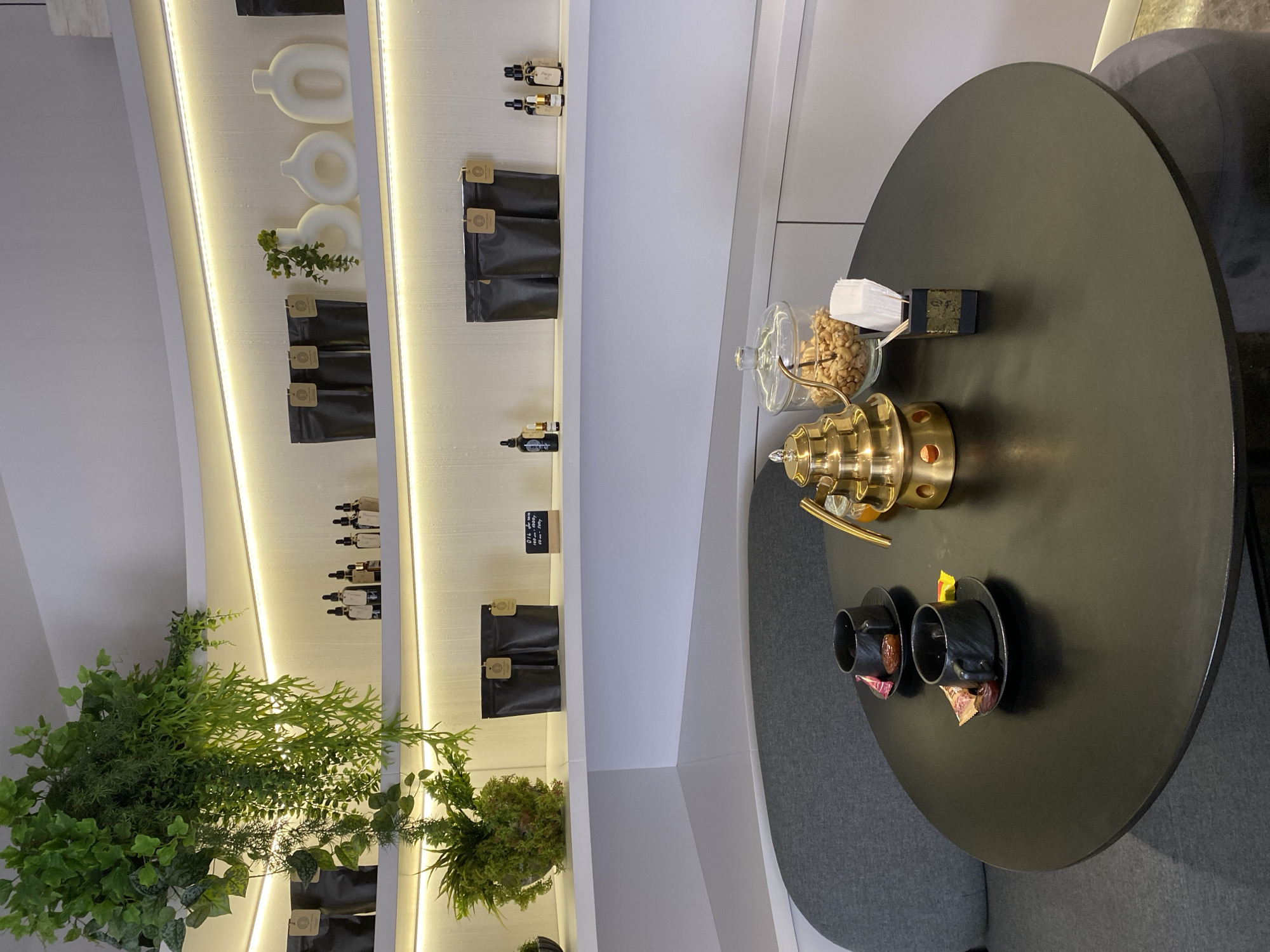 The image shows a modern interior, potentially a shop or café, though no people are visible. A brightly lit shelving unit displays various products, including what appear to be packages of dried goods, vials of liquids, and decorative ceramic items. Artificial plants are also interspersed among the products. To the right, a round black table is set for tea or coffee service. It features a brass teapot, two black cups, a bowl of snacks covered by a glass lid, and a stack of white napkins. The chairs around the table are upholstered in gray fabric. The precise location within Votkinsk, Russia, cannot be determined from the visual information provided.