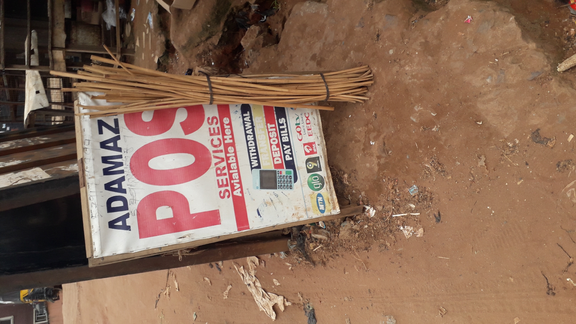 A roadside advertising board for "ADAMAZ POS SERVICES" is leaning against a dark structure. The sign displays "POS SERVICES Available Here" in large red letters. Below this, it lists services such as "WITHDRAWAL," "TRANSFER," "DEPOSIT," and "PAY BILLS," accompanied by images of a POS device and logos for MTN, Glo, Airtel, and EEDC. A bundle of dry sticks is tied to the right side of the advertising board. The ground surrounding the sign is uneven, dusty, and littered with debris, including plastic bags and other refuse. In the background, parts of market stalls and a yellow vehicle are visible. The overall scene depicts a typical informal business setup in an outdoor urban environment.