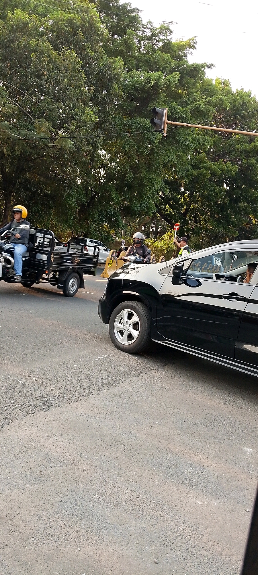 This is a ground-level, vertical-format, outdoor photograph taken from a slightly elevated perspective on a paved road, likely in Depok, Indonesia. The scene captures a traffic intersection with vehicles and pedestrians.
In the foreground, the right side of a black SUV is prominently visible, reflecting the bright, overcast sky and surrounding trees. The SUV has silver alloy wheels and a reflective silver strip along its lower body. Inside the SUV, a person can be seen in the driver's seat, wearing glasses.
To the left of the SUV, a three-wheeled vehicle, possibly a motor tricycle or "bajaj," is also on the road. It is black with a large cargo bed. The driver, wearing a yellow helmet and a grey jacket, is looking ahead. A second person, wearing a grey helmet and jacket, is riding behind the driver on the same three-wheeled vehicle, also looking forward.
In the background, there are several other vehicles, including a white car, and a traffic signal pole with two dark traffic lights. A person in a vest is holding a "STOP" sign, indicating traffic control. Lush green trees with dense foliage form a backdrop for the scene, suggesting a location with significant vegetation, consistent with Depok's environment. The overall lighting indicates it is daytime, possibly late afternoon due to the warm tones. The road surface appears to be asphalt with visible texture and some faint white markings.