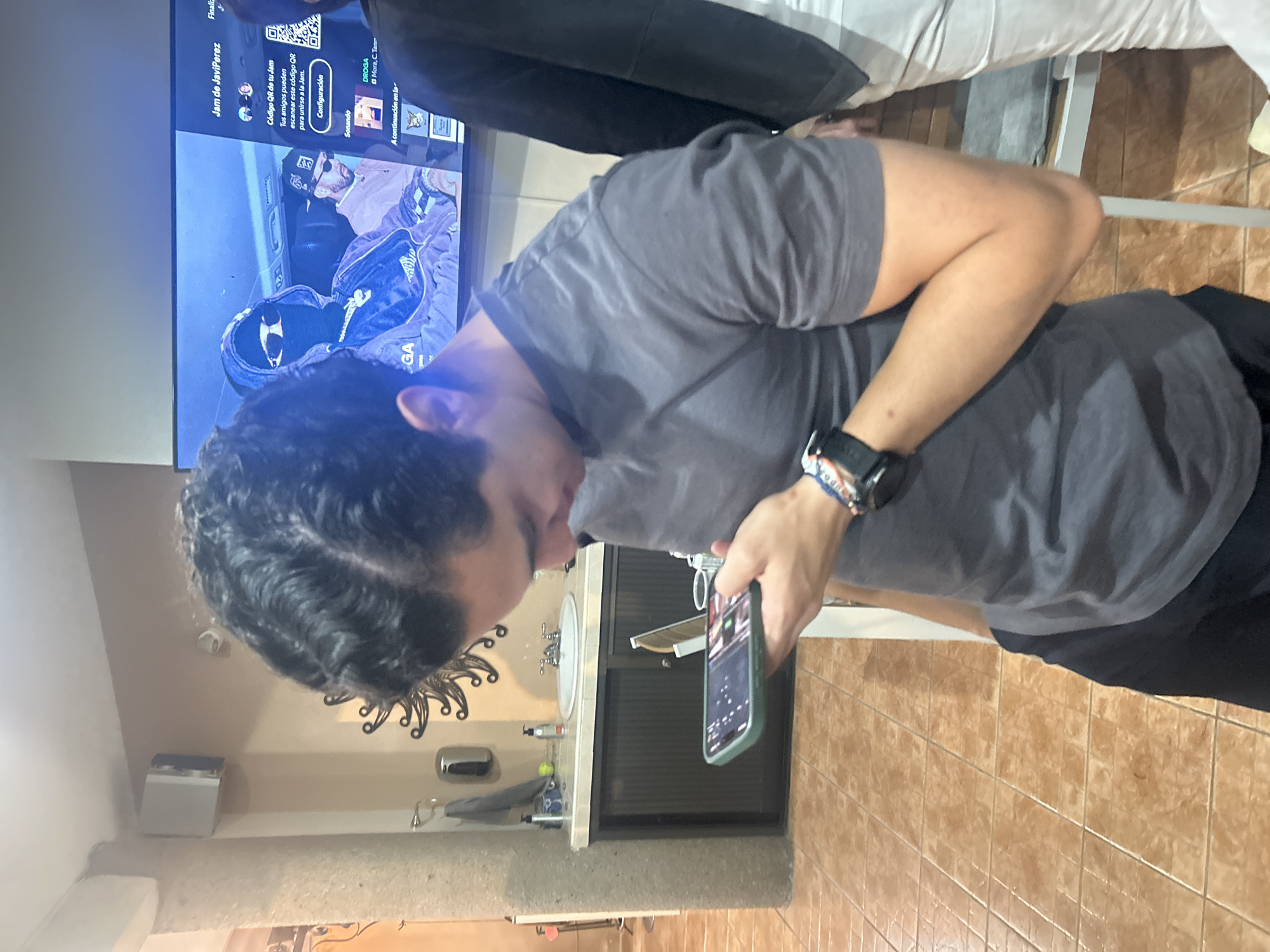 A person in a gray t-shirt is holding a smartphone in their right hand, looking down at the screen. The phone has a green case. The person is standing in a bathroom with brown tile flooring. Behind the person is a wall with a sink, a decorative wall piece, and bathroom accessories. In the background, a television screen displays a "Jam de JaviPerez" interface, complete with a QR code and song information. The location is San Juan Cuautlancingo, Mexico.