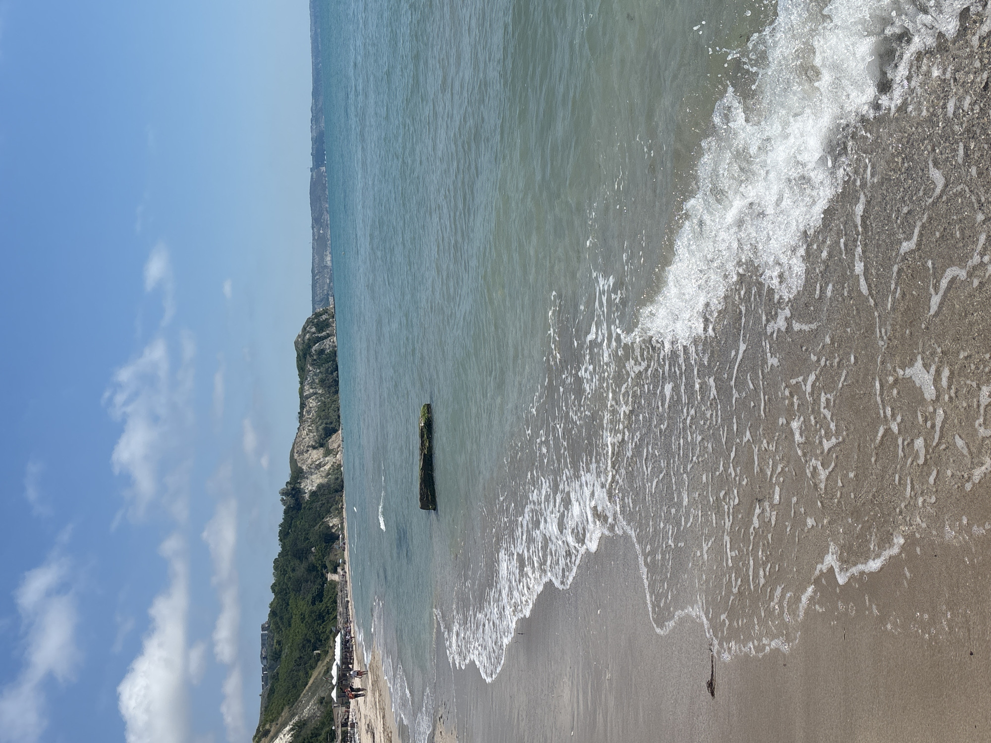 The image shows a beach scene in Topola, Bulgaria, on a sunny day. The foreground is dominated by the sandy beach with gentle waves lapping at the shore. The water is a clear turquoise color. In the middle ground, there is a large, rectangular piece of driftwood or debris partially submerged in the water, covered in green algae. Along the coastline, cliffs covered in green vegetation rise up, with some buildings visible on the top of the cliffs in the distance. There are several white umbrellas and lounge chairs set up on the beach, indicating a resort or a public beach area. A few people are walking along the shoreline. The sky is blue with scattered white clouds. The overall impression is a serene and picturesque coastal landscape.