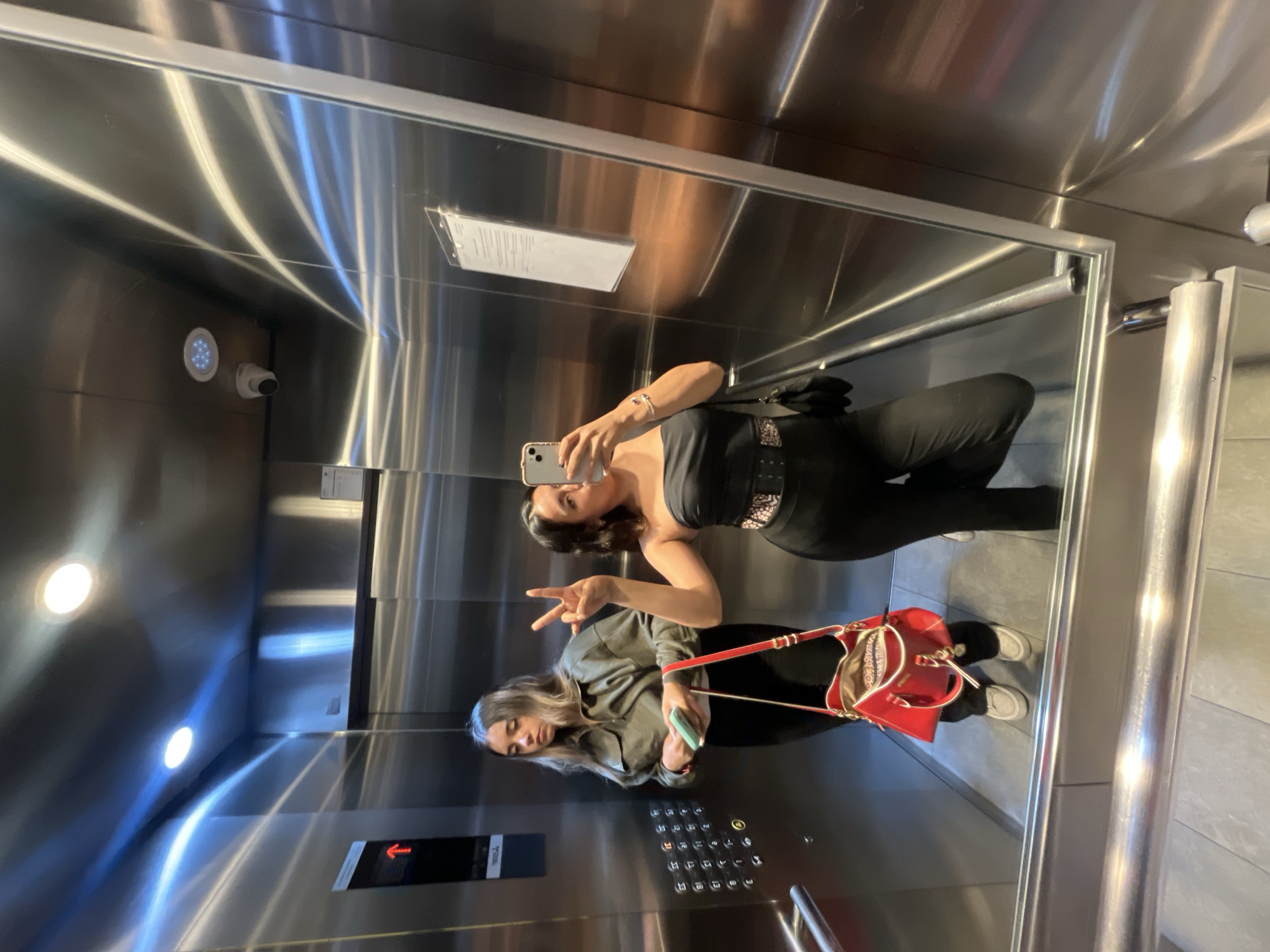 Two women are taking a selfie in an elevator. The elevator has a stainless steel interior, with a mirror on one wall. One woman is holding a smartphone and posing. The other woman is making a peace sign. Both women are wearing clothing that appears dark. One has a red purse with her. Elevator buttons are visible. The location is Unknown, Unknown.