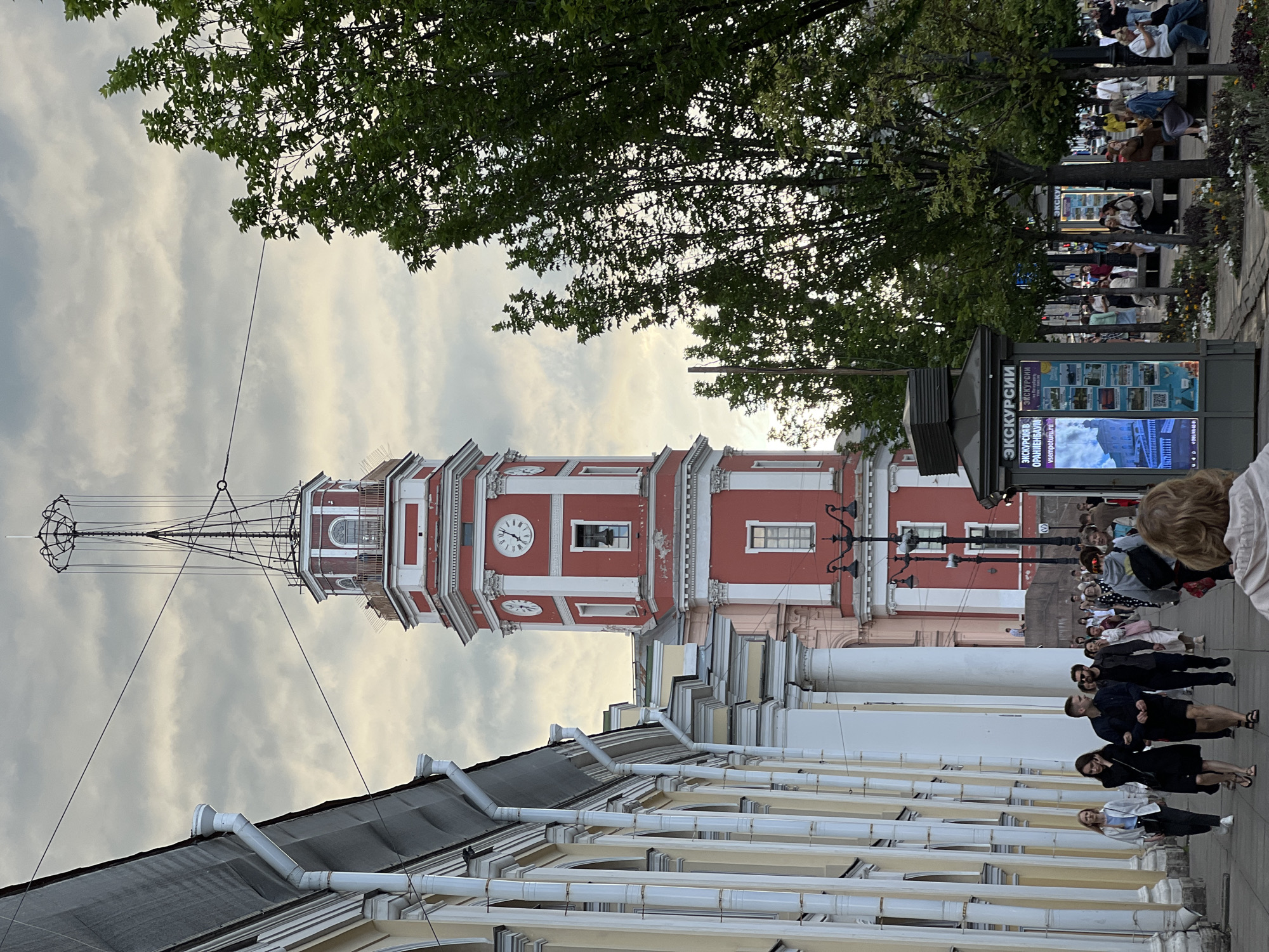 The image depicts the Peter and Paul Fortress Clock Tower, a prominent landmark in Saint Petersburg, Russia, against a cloudy sky. The tower, painted in red and white, features clock faces and a bell. To the left of the tower is a building with a colonnade facade, painted in pale yellow and white. In the foreground, a paved walkway is lined with trees and people are walking and sitting. Some individuals are captured in motion, while others are interacting or observing. A kiosk with information about excursions is also visible. The overall atmosphere suggests a moderately busy urban environment.