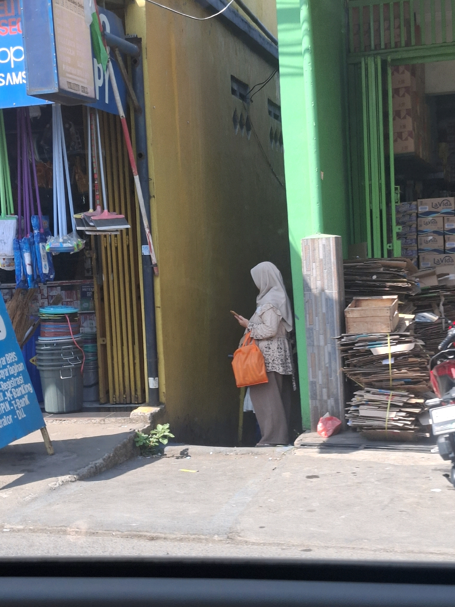 A woman wearing a light-colored hijab and a patterned top is standing near a building. She is holding a smartphone in her right hand and has an orange tote bag on her left arm. She is positioned between a yellow building on her left and a green column on her right. To the right of the green column, there is a stack of cardboard boxes and a red motorcycle. In the background, there is an open doorway with green metal gates, revealing more boxes inside. In front of the yellow building, several cleaning supplies, including brooms and mops, are displayed. A blue sign with white text is leaning against the building on the left. The ground is paved and there is some vegetation growing in a crack in the pavement. The scene appears to be an outdoor area in front of a shop or market.